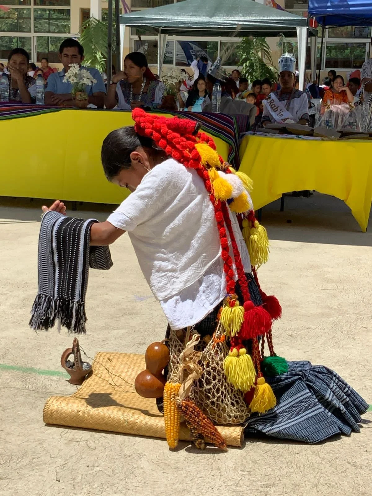 Una mujer en ropa tradicional indígena en una ceremonia o festival, inclinada en una postura de ofrenda o respeto, rodeada de objetos culturales y dispuesta en una superficie de tela, con un grupo de personas observando en el fondo.