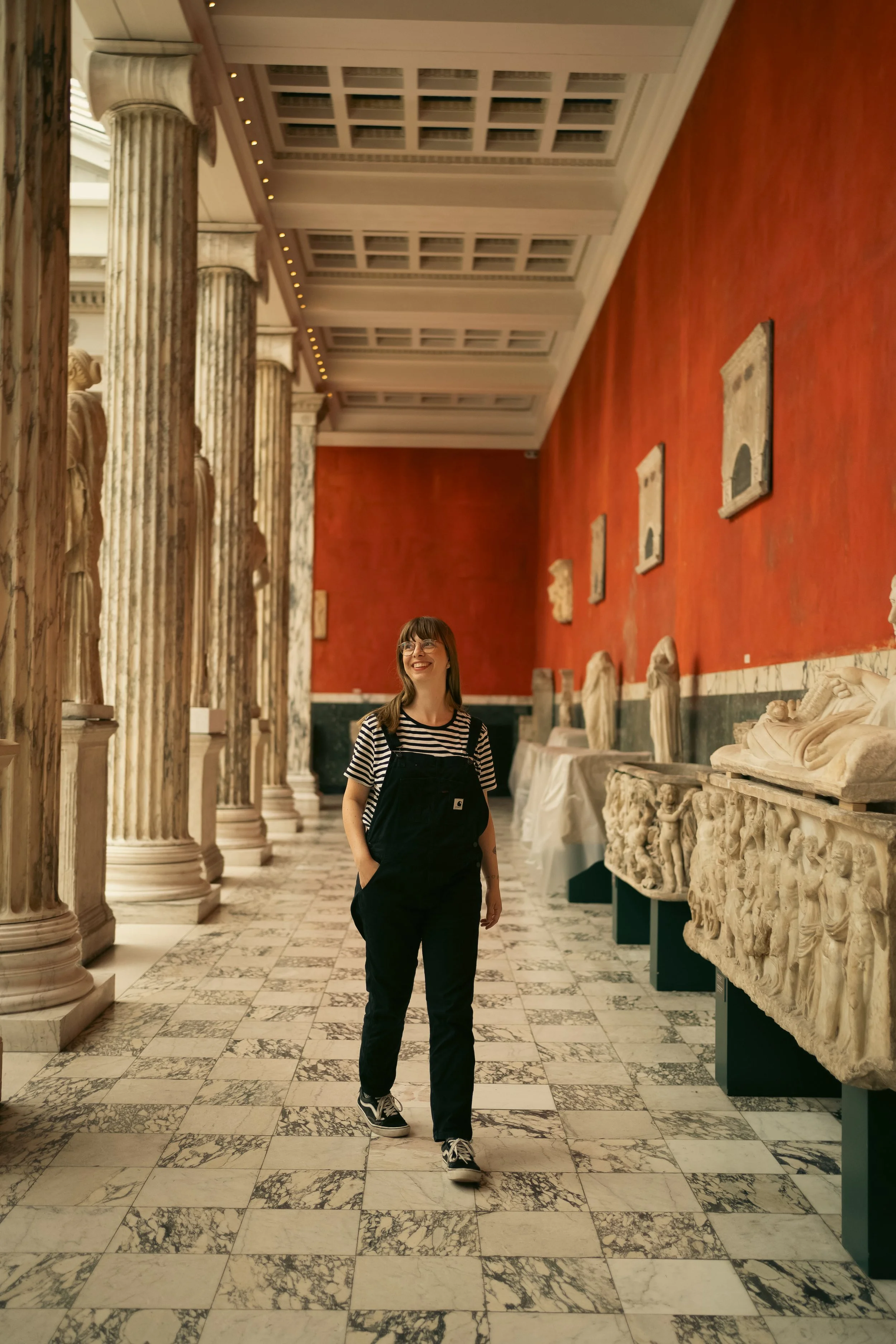 A woman walking in a museum hallway with ancient Greek or Roman sculptures and artifacts, featuring marble columns and carvings, and a red wall in the background.