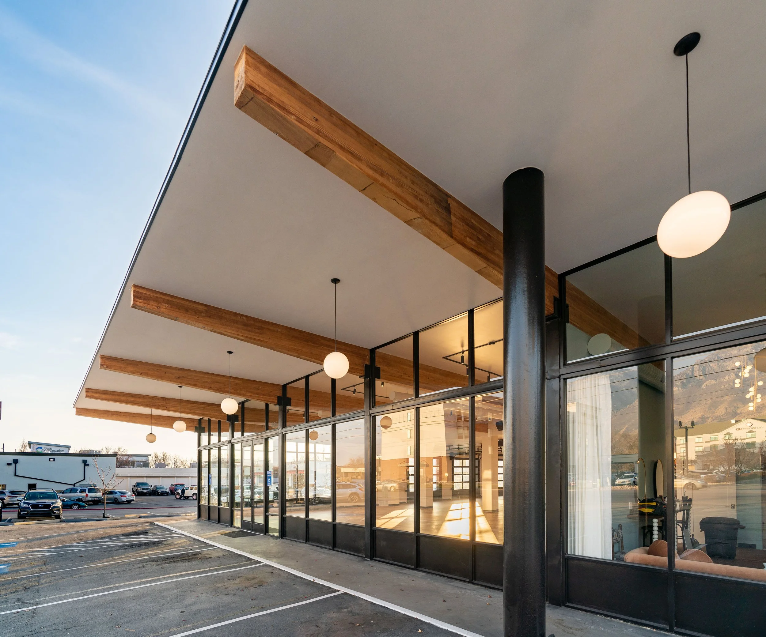 A modern building with large glass windows, wooden beams on the ceiling, and hanging white round lights, with a parking lot and cars outside.