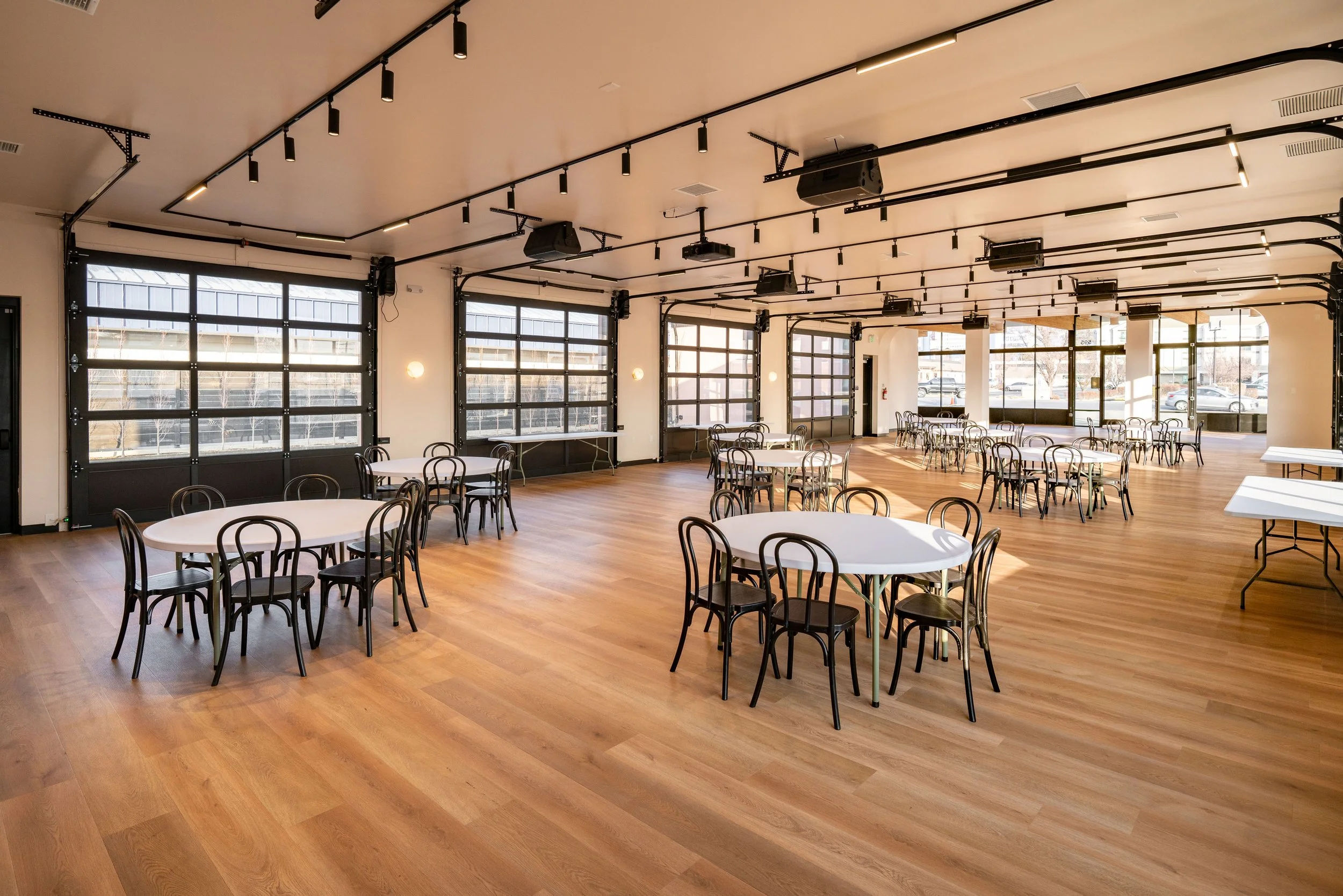 Spacious event hall with wooden floors, large windows, round tables with black chairs, and ceiling-mounted black lighting and speaker systems.