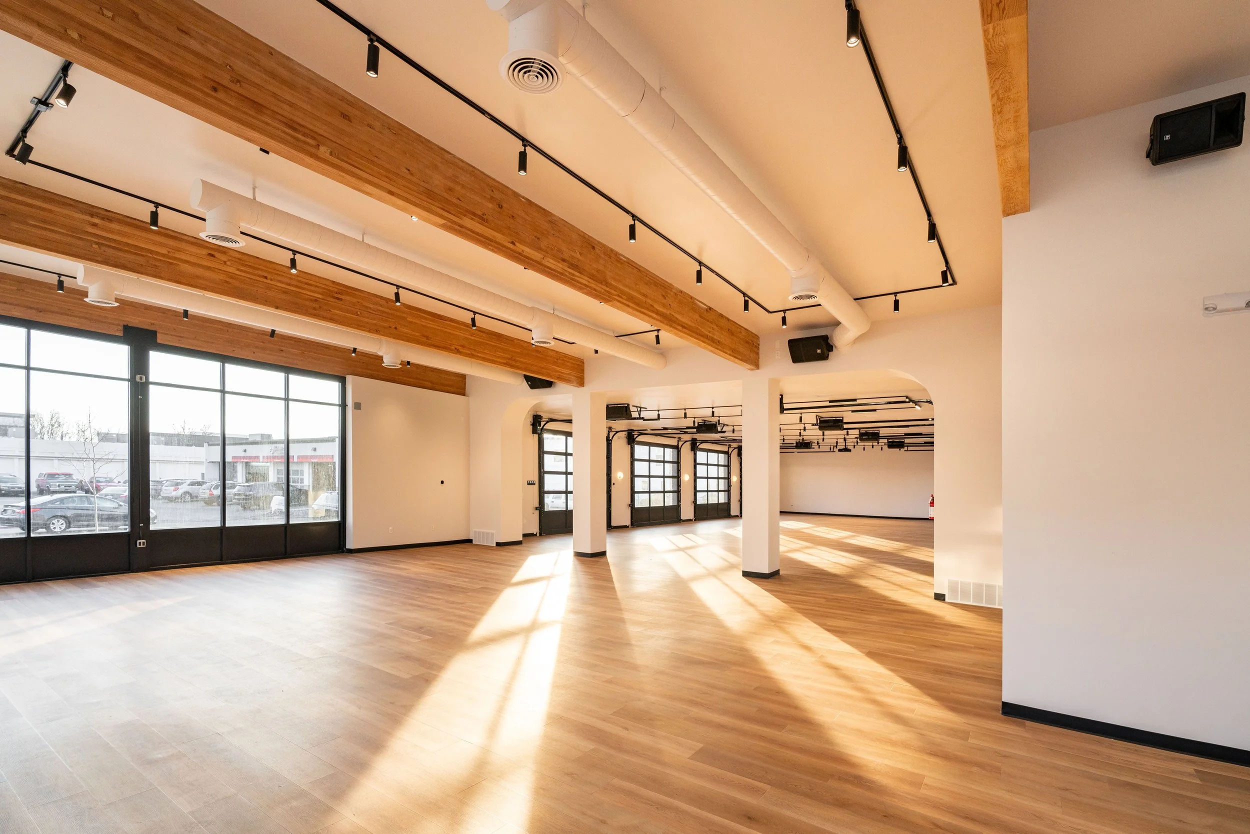 An empty room with large windows, hardwood floors, exposed ceiling beams, and modern lighting.