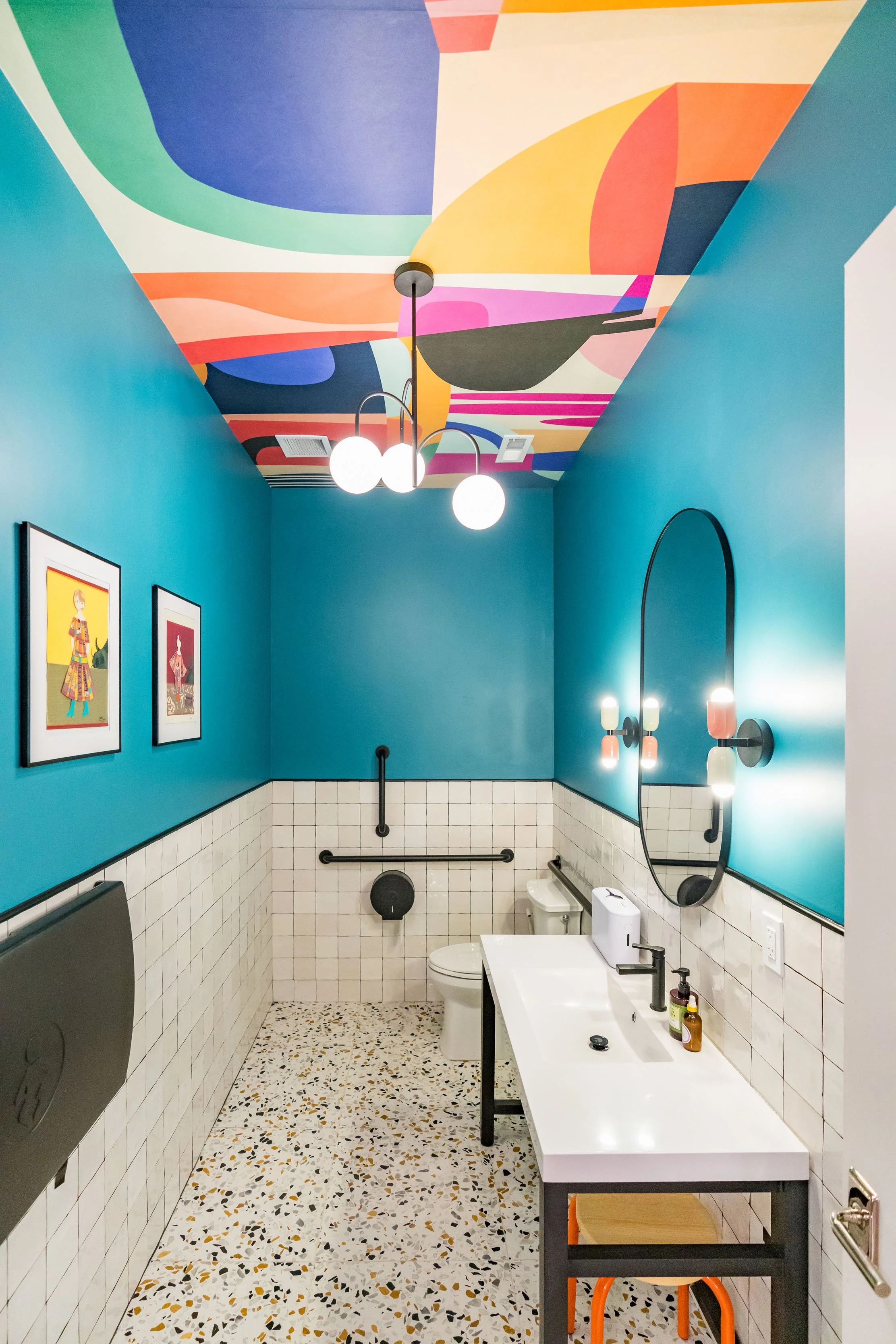 A colorful bathroom with a vibrant abstract painted ceiling, turquoise walls, white tiled lower walls, a white sink with black fixtures, an oval mirror, framed artwork on the wall, and a toilet with back support rails. The bathroom floor has a terraz