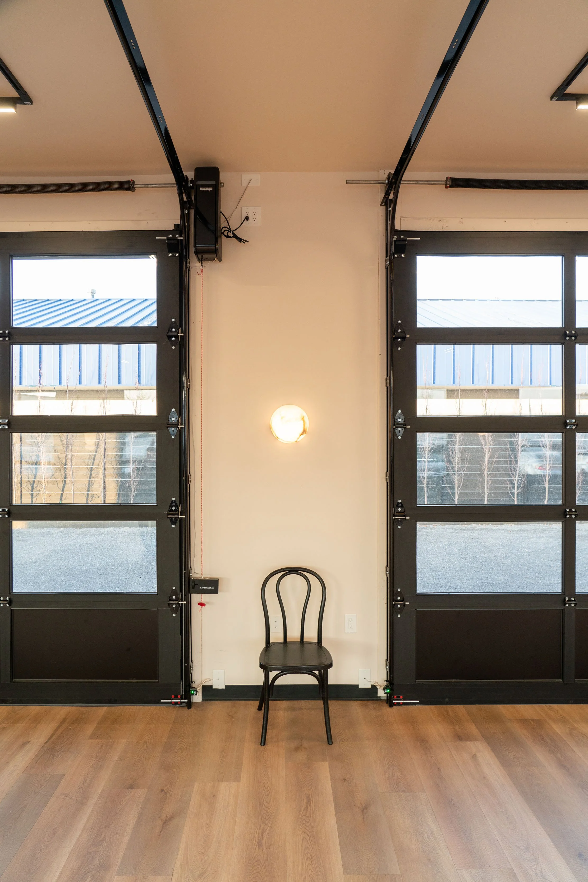 Interior of a garage with two black garage doors, a black chair in the middle, a small wall-mounted light fixture, and a woven wood wall slightly visible in the background.