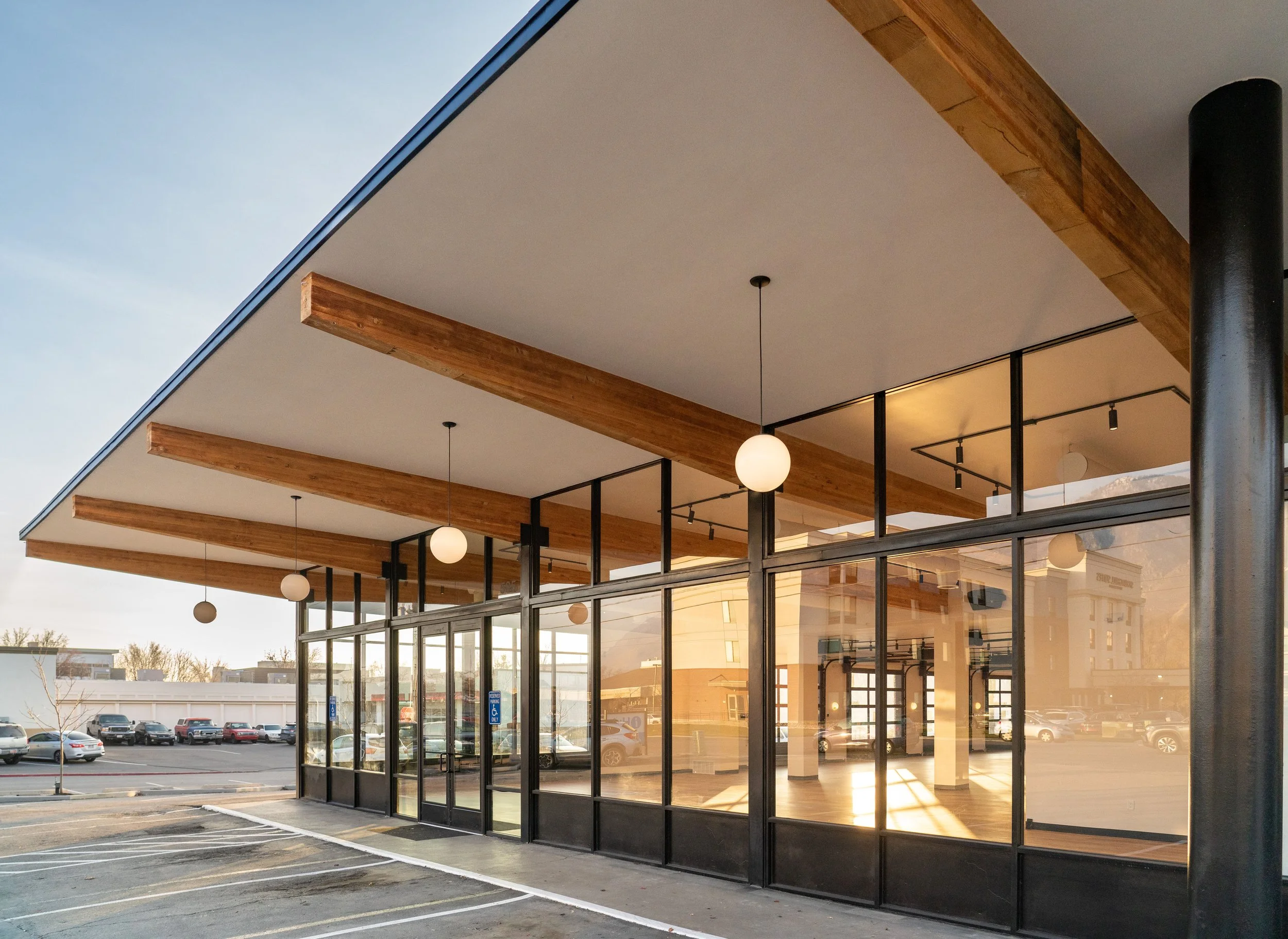 Modern commercial building with large glass windows, wooden beams on the ceiling, and hanging spherical lights, situated in a parking lot during sunset.