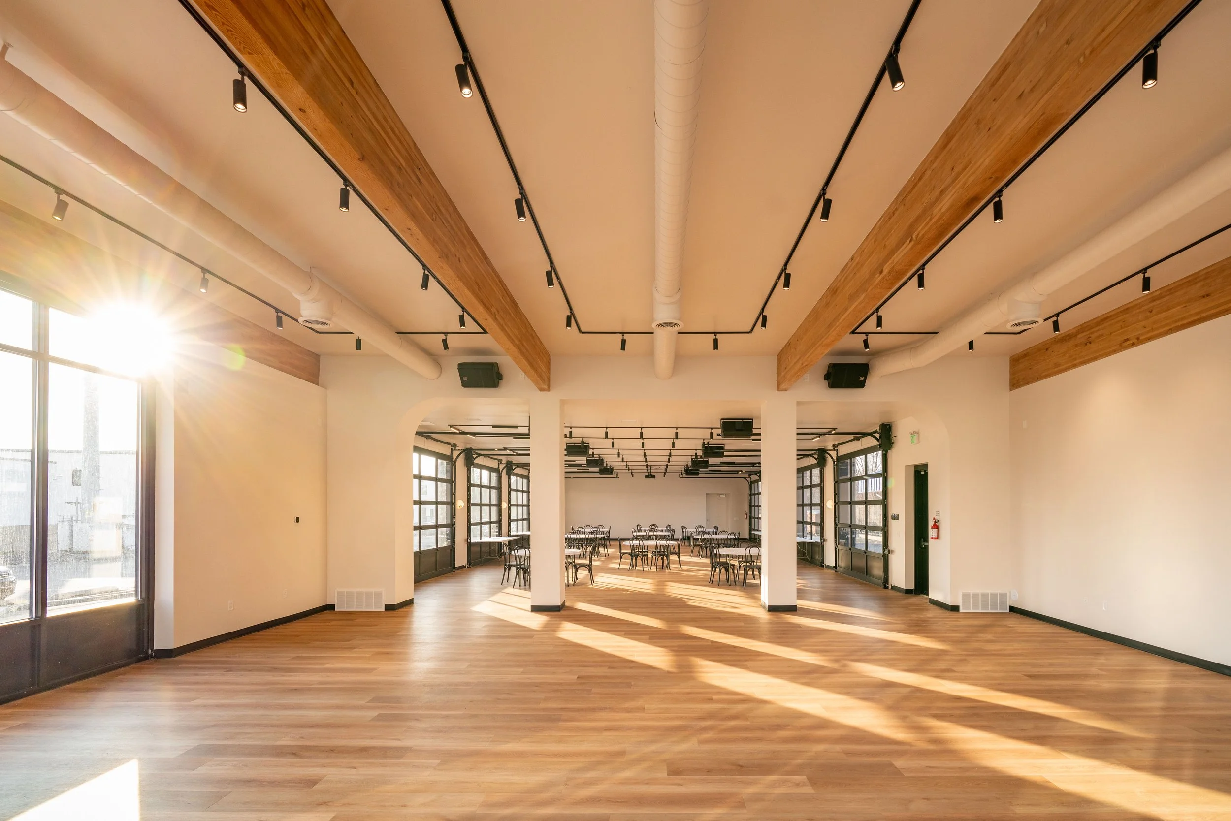 Bright, spacious event hall with wooden floors, large windows, and white walls. The ceiling has wooden beams and black track lighting. Several tables and chairs are arranged towards the back, with sunlight streaming through the windows.