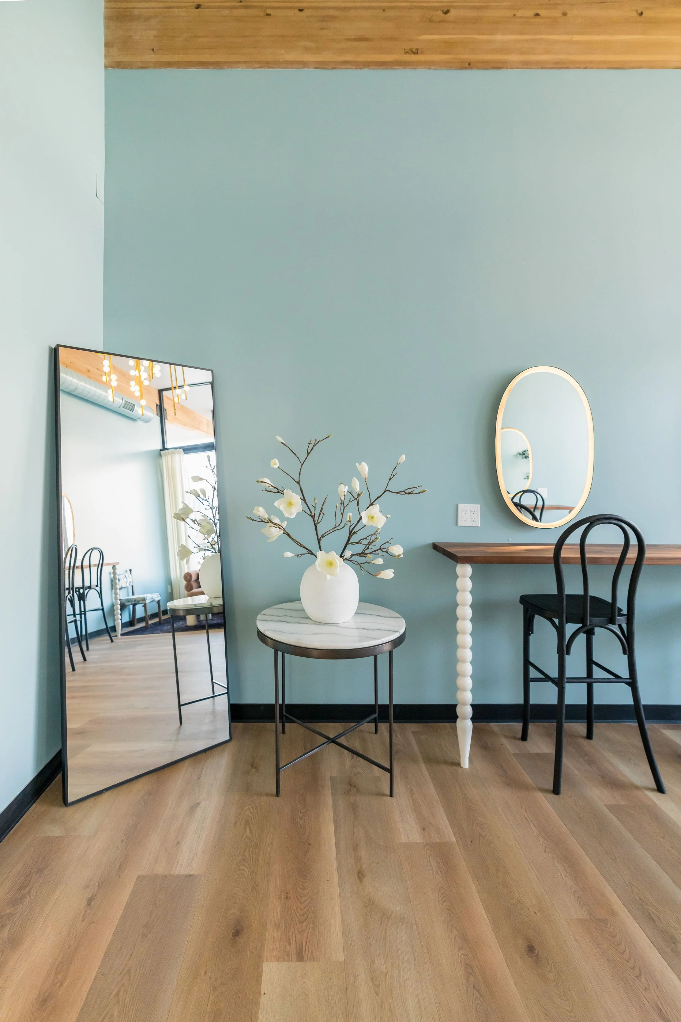 Interior of a room with light blue walls, a large mirror leaning against the wall, a small round table with a white vase and flower branch, a wooden desk with a black chair, and a gold-framed oval mirror above the desk.