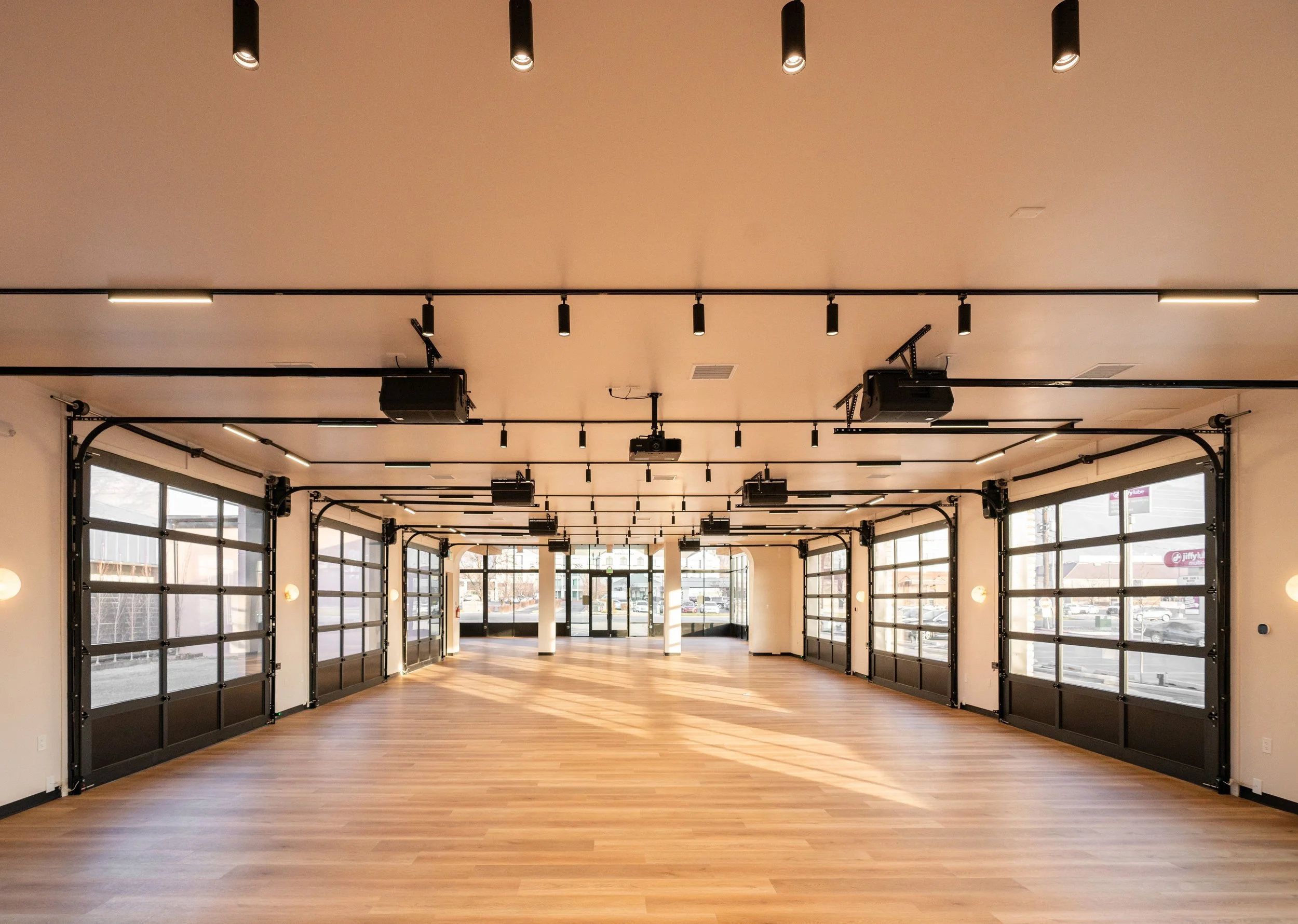 Empty spacious commercial space with large garage-style glass doors and hardwood floors, illuminated by ceiling lights, with natural light coming through the windows.