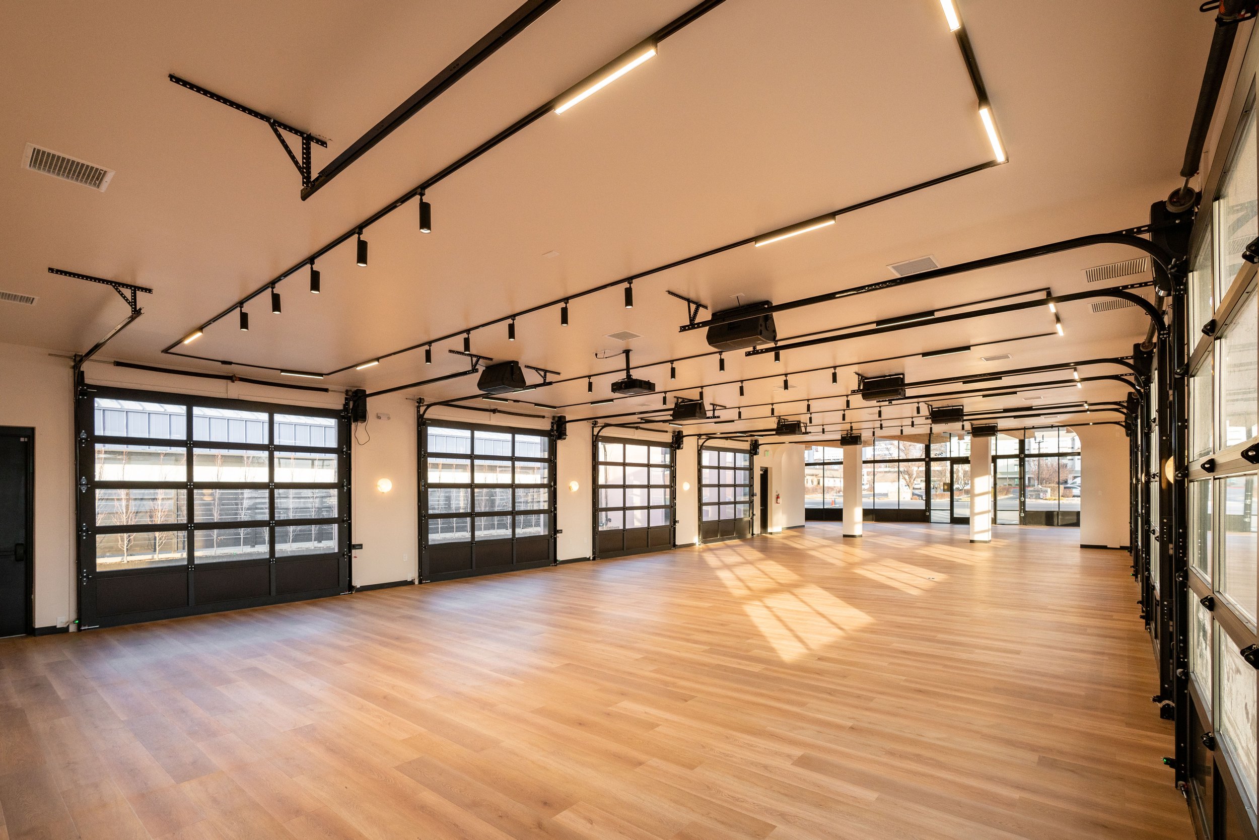 Empty indoor space with wooden flooring, large windows, and garage-style doors, equipped for fitness or events.