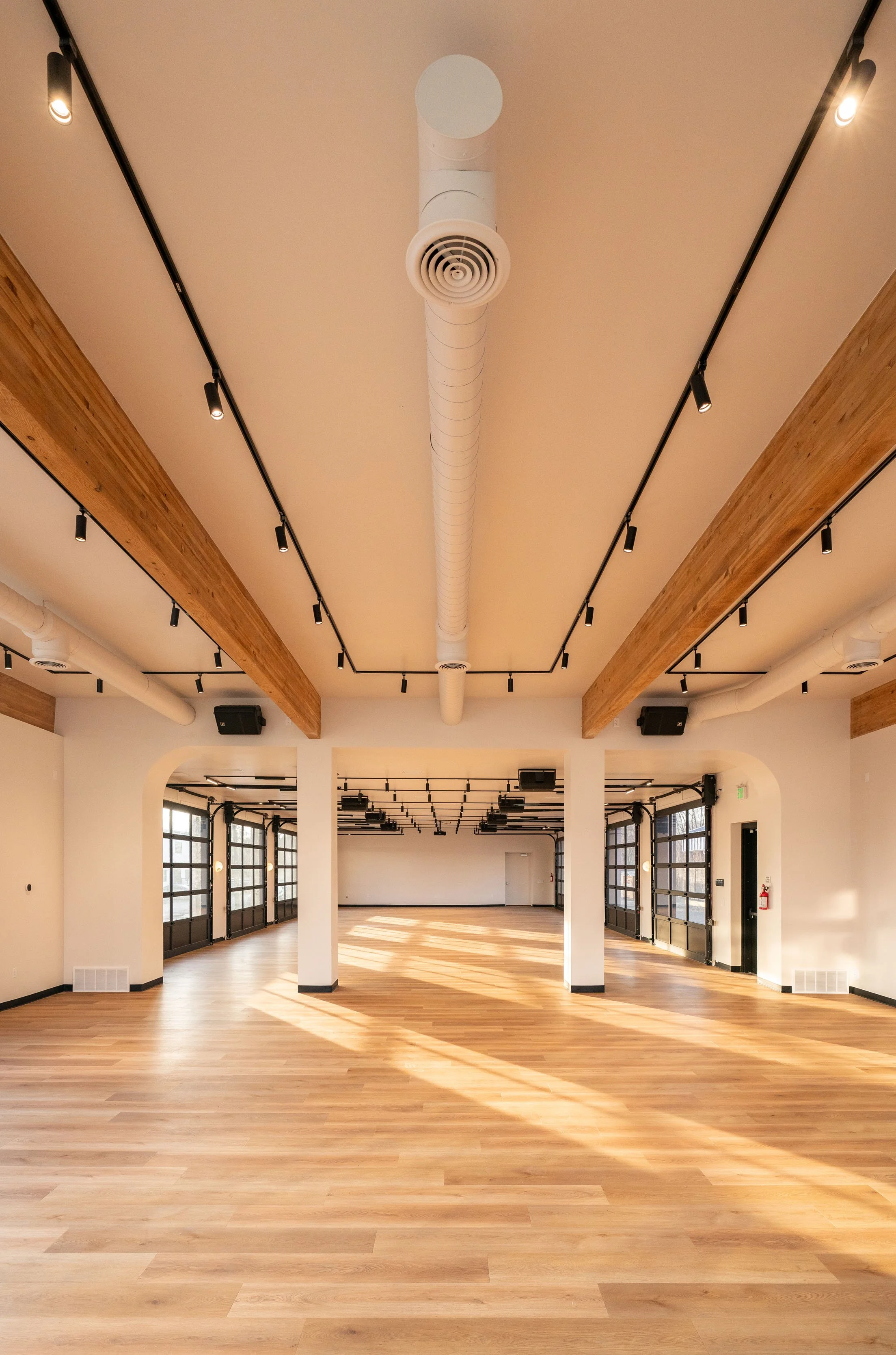 Bright, empty modern event space with wooden floors, large sliding glass doors, exposed wooden beams, white walls, and track lighting.