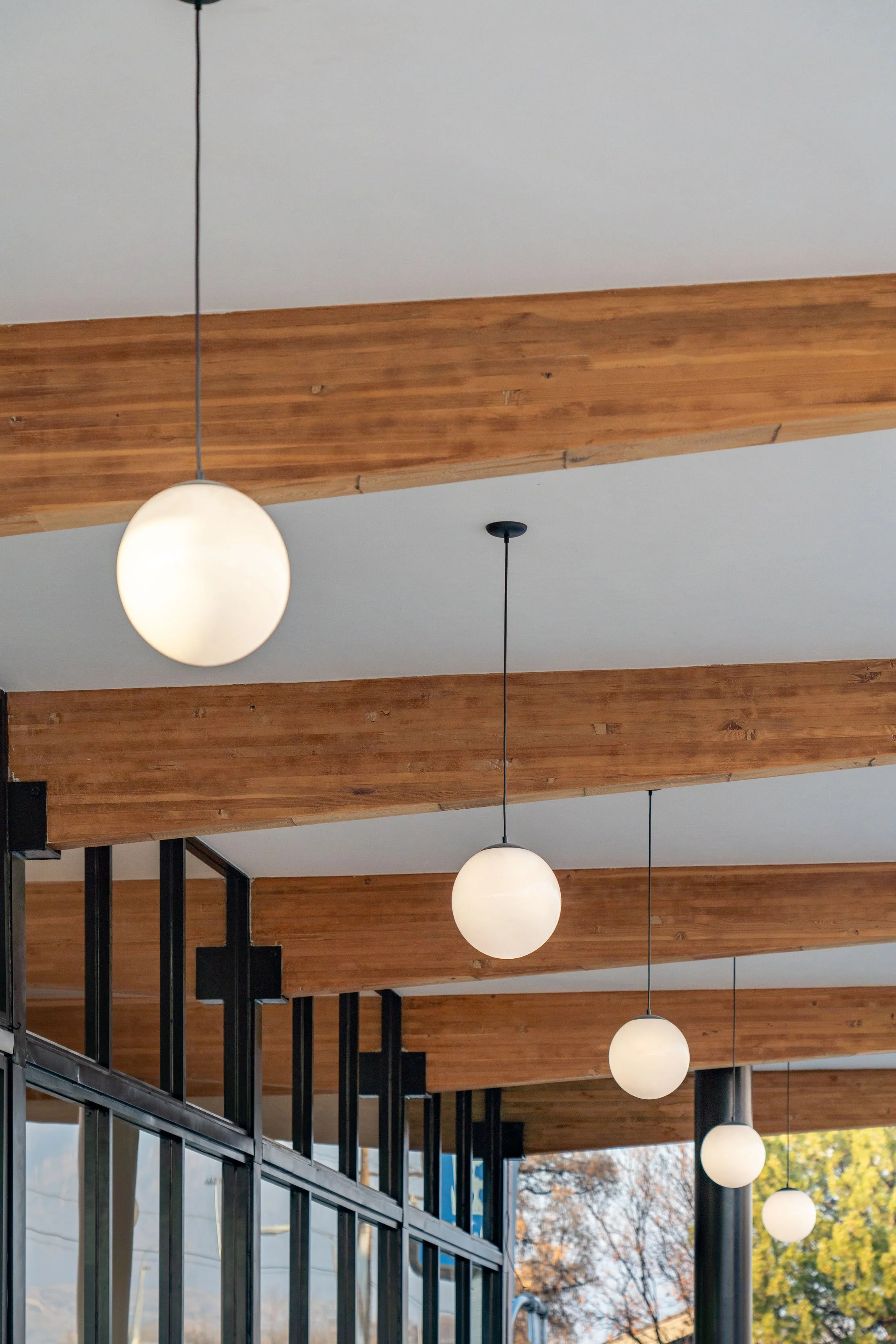 Interior view of a modern building with wooden beams on the ceiling and hanging spherical pendant lights. Large windows with black frames are visible, showing trees outside.
