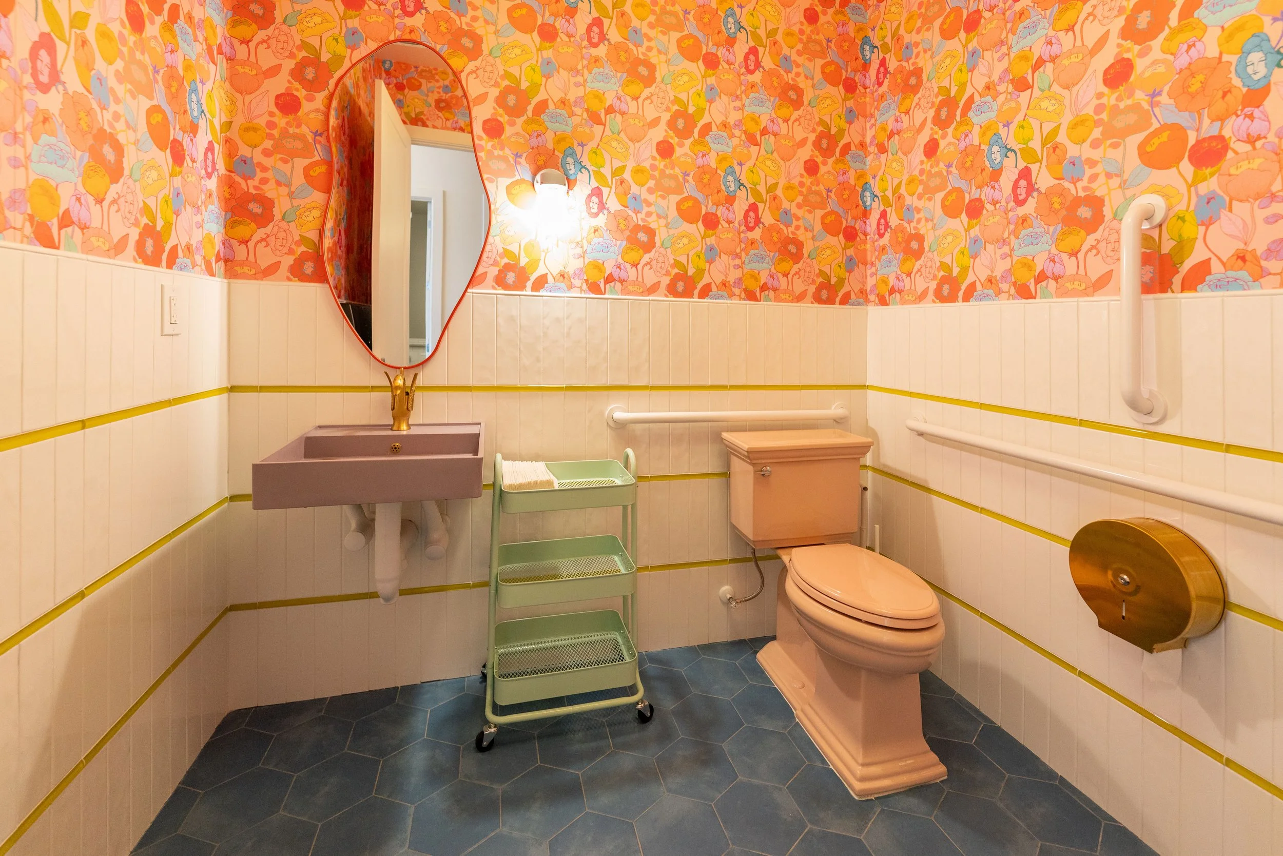 Bathroom with colorful floral wallpaper, pink toilet, pink sink with gold faucet, oval mirror, white wall paneling with yellow trim, small green storage cart, blue hexagonal tile flooring, white safety bars and brown tissue holder.