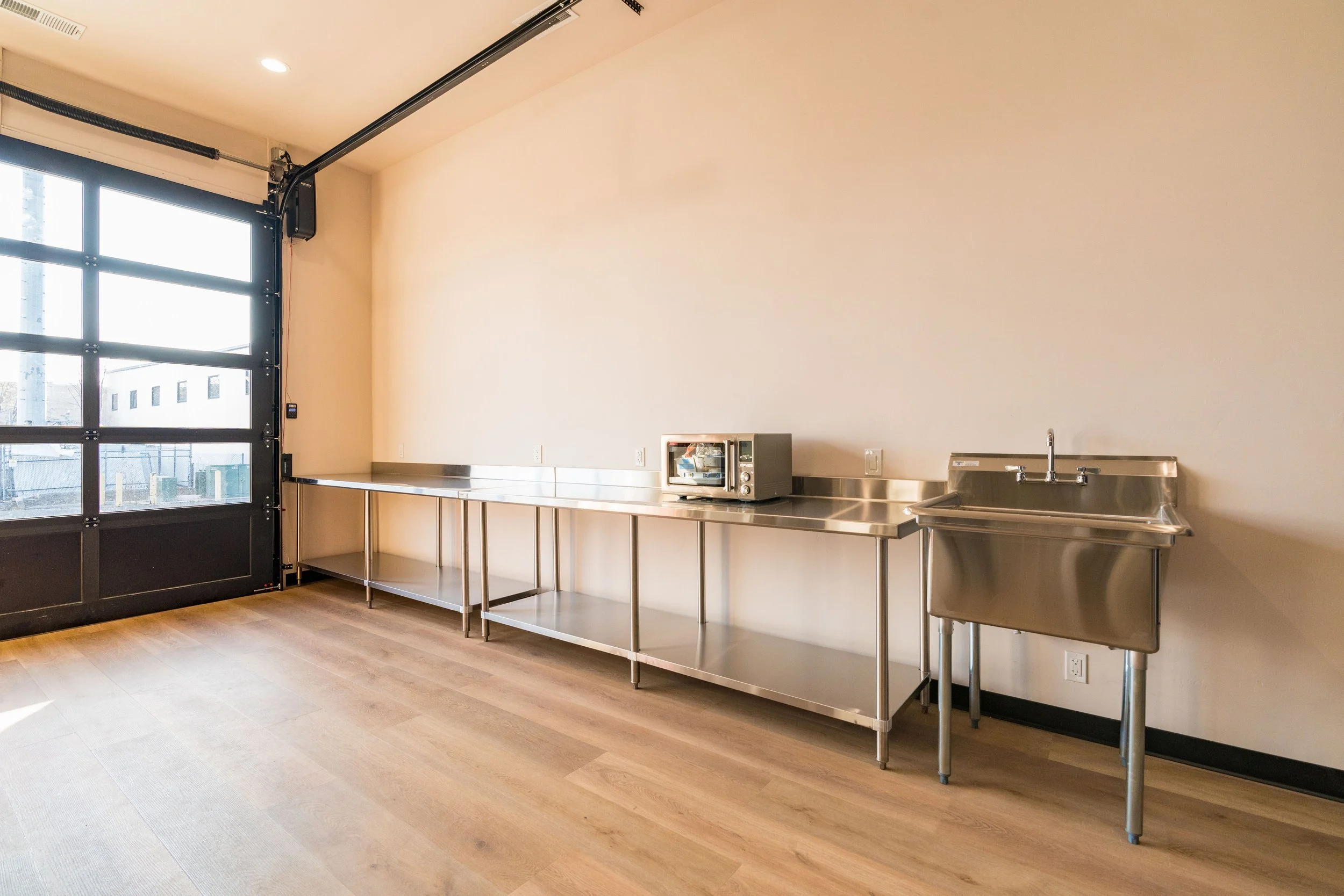 Empty commercial kitchen with stainless steel counters, a microwave, and a utility sink in a room with wood flooring and a large glass garage door.