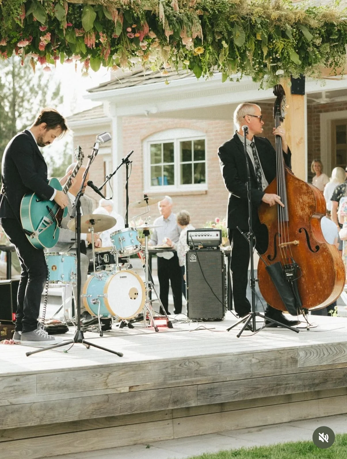 A band performing live on an outdoor wooden stage at a social gathering, with a guitarist, double bassist, and drummer, under a floral canopy.