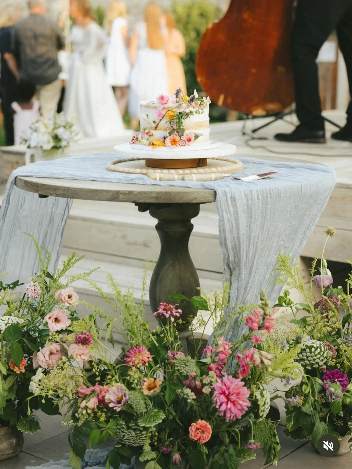 A small, unfinished wedding cake decorated with fresh flowers on a table with a white cloth at an outdoor reception. There are colorful flower arrangements surrounding the table.