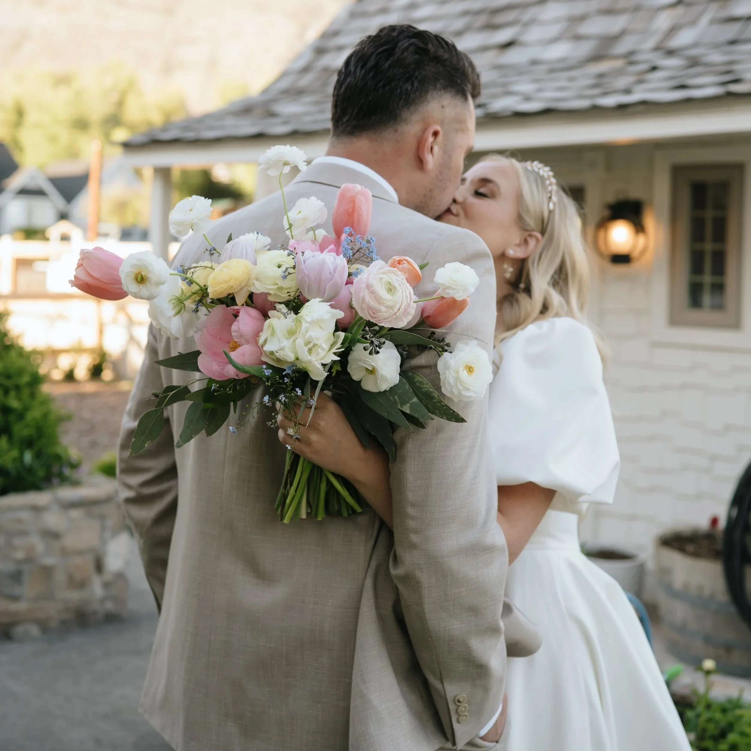 A bride holding a bouquet of pink and white flowers, wearing a white wedding dress with off-the-shoulder sleeves and sheer fabric, standing outdoors.