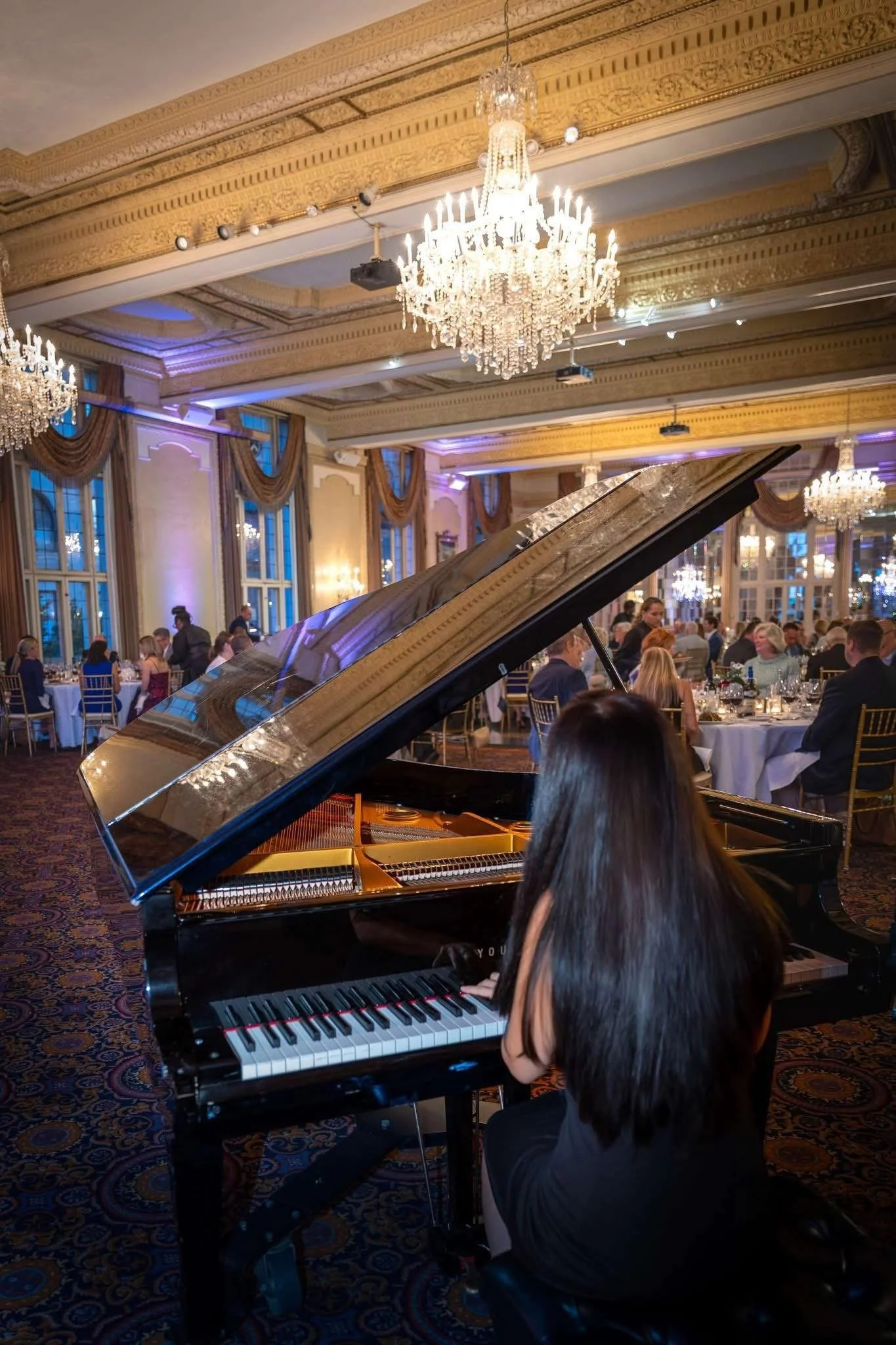 A woman playing a grand piano at a formal event in an ornate ballroom with chandeliers and large windows.