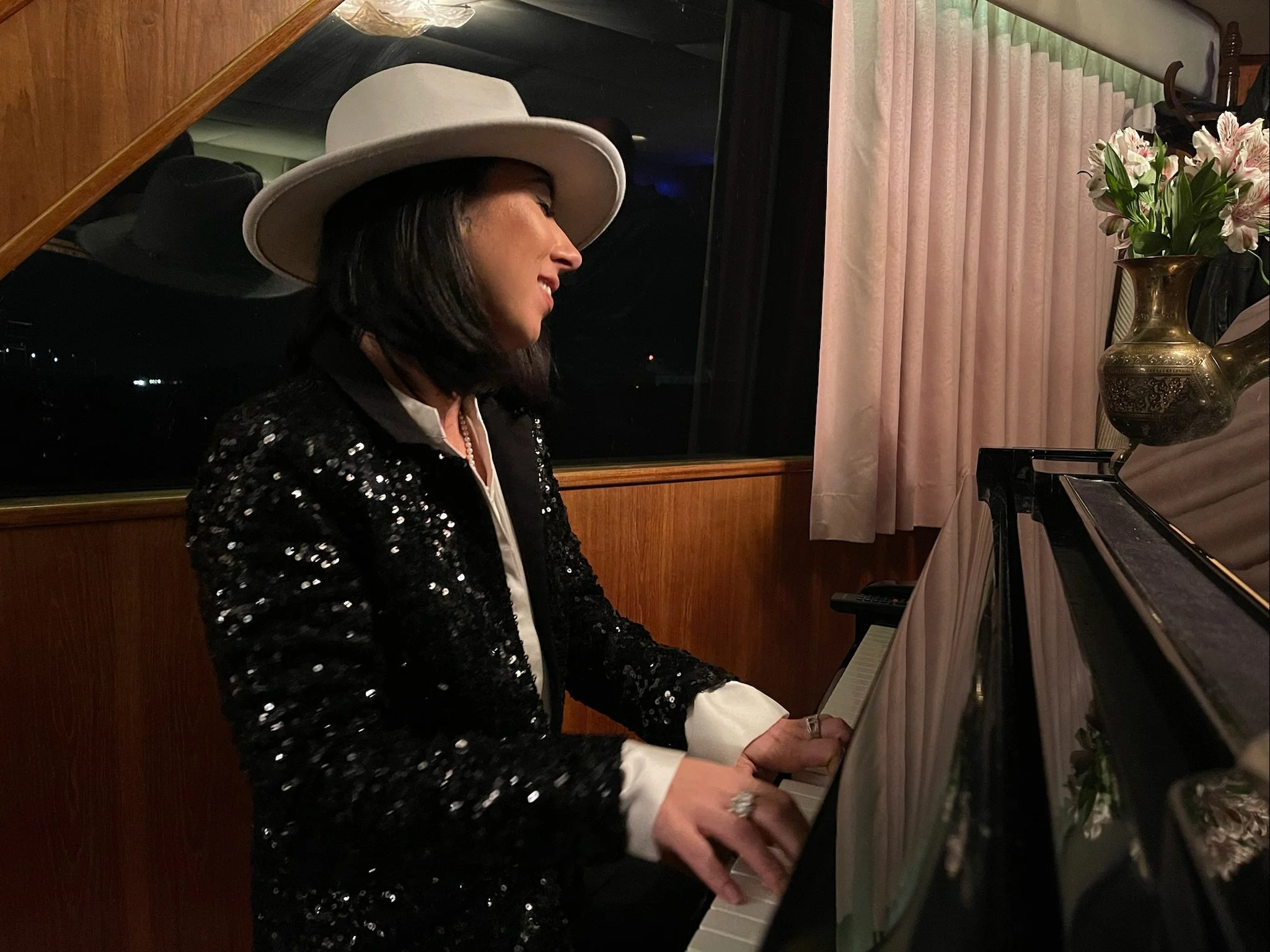 A woman wearing a white hat and a black sequined jacket is playing a piano in a cozy, dimly-lit room. She has shoulder-length dark hair and is smiling softly. There is a vase of flowers on the piano and a window with light-colored curtains in the background.