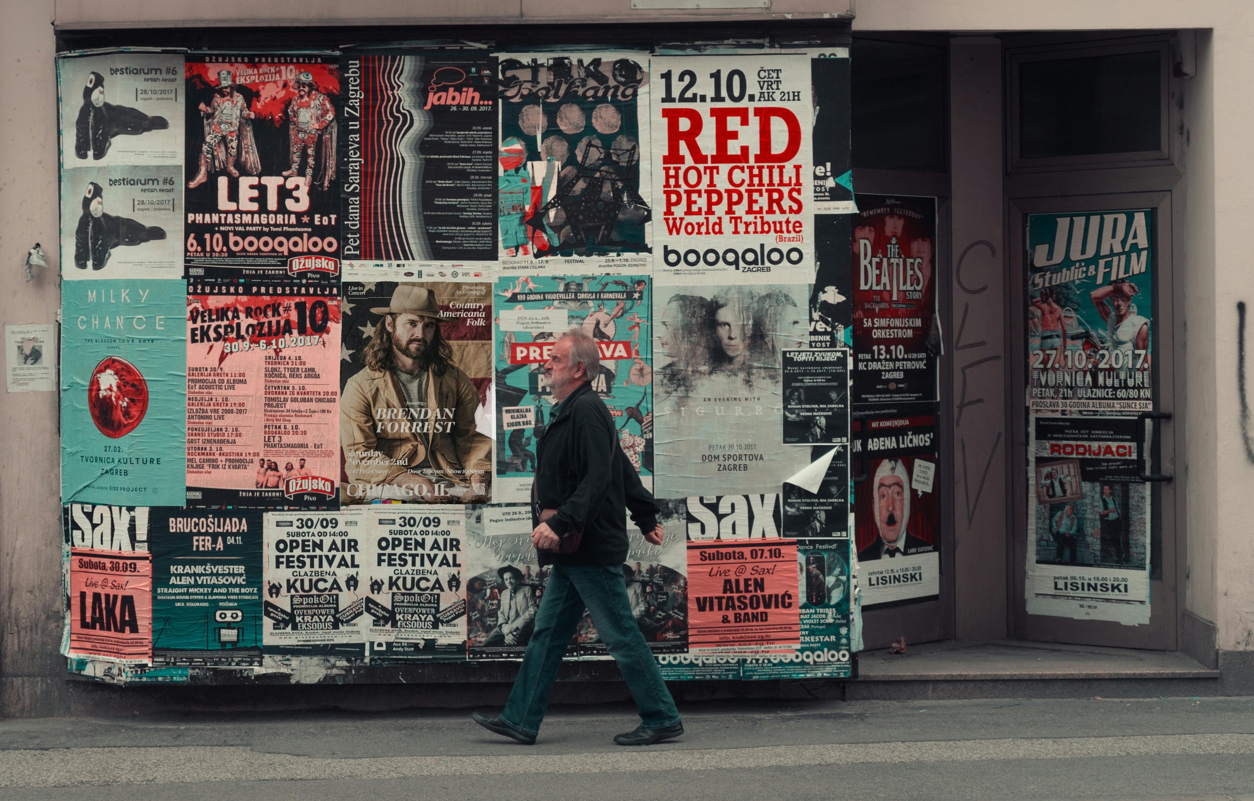 A man with white hair and beard walking past a large billboard covered in colorful concert and event posters on a city street.
