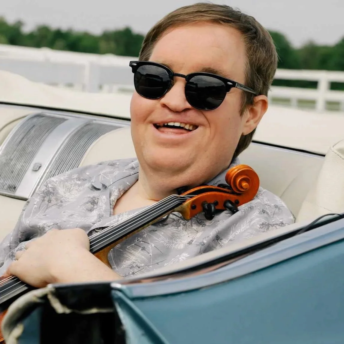 Grammy Award Winner Michael Cleveland, smiling and wearing sunglasses, plays the violin while seated in a white interior car.