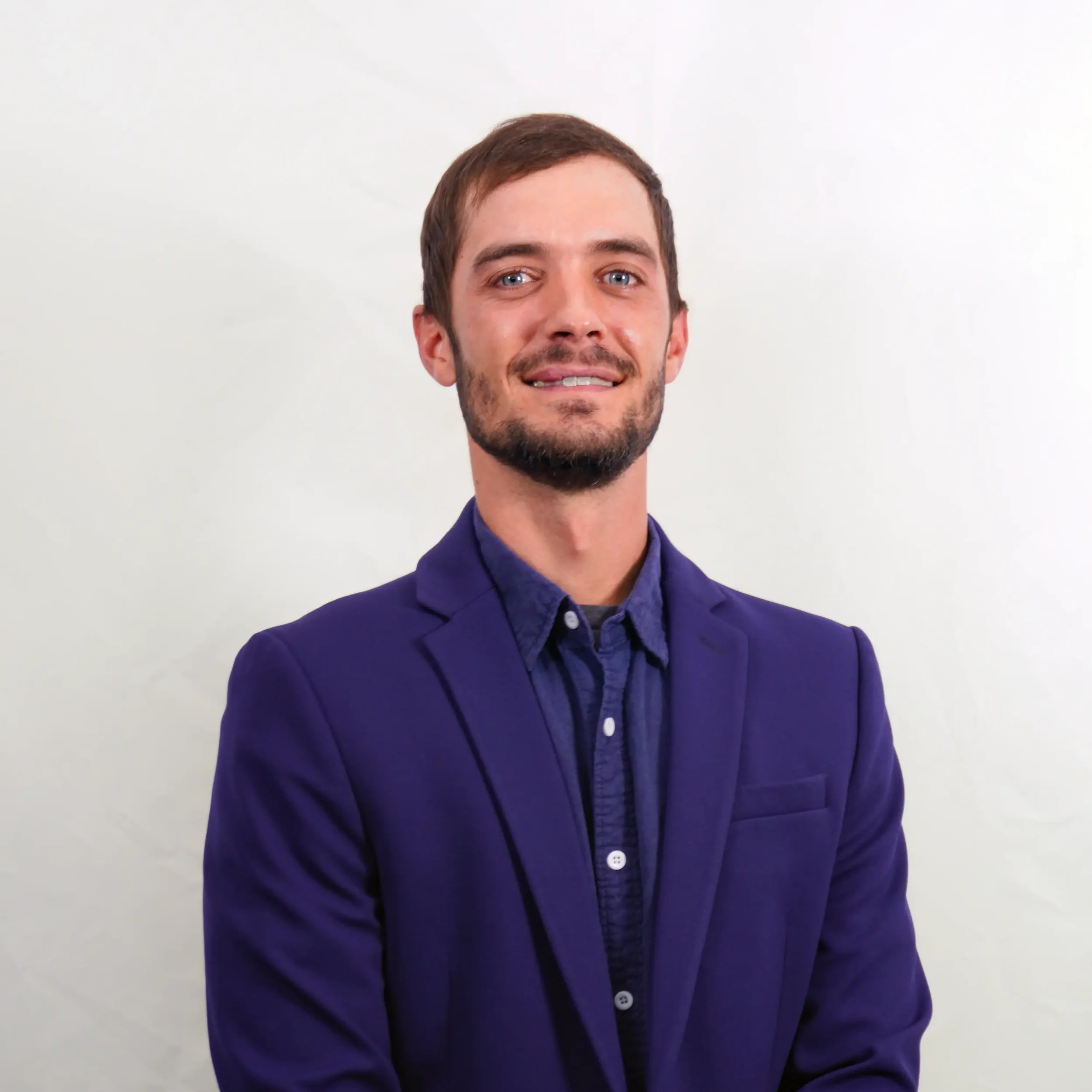 A young man with brown hair and a beard, wearing a plaid shirt, smiling at the camera against a plain white background.