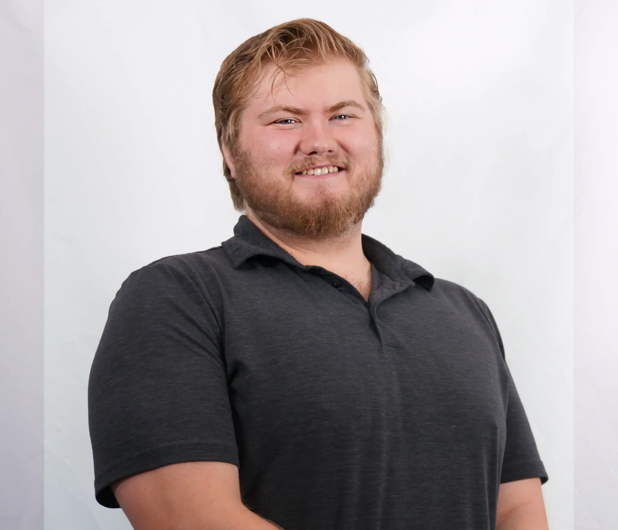 A young man with blonde hair, light beard, and mustache, wearing a black collared shirt, smiling and looking at the camera against a plain light-colored background.