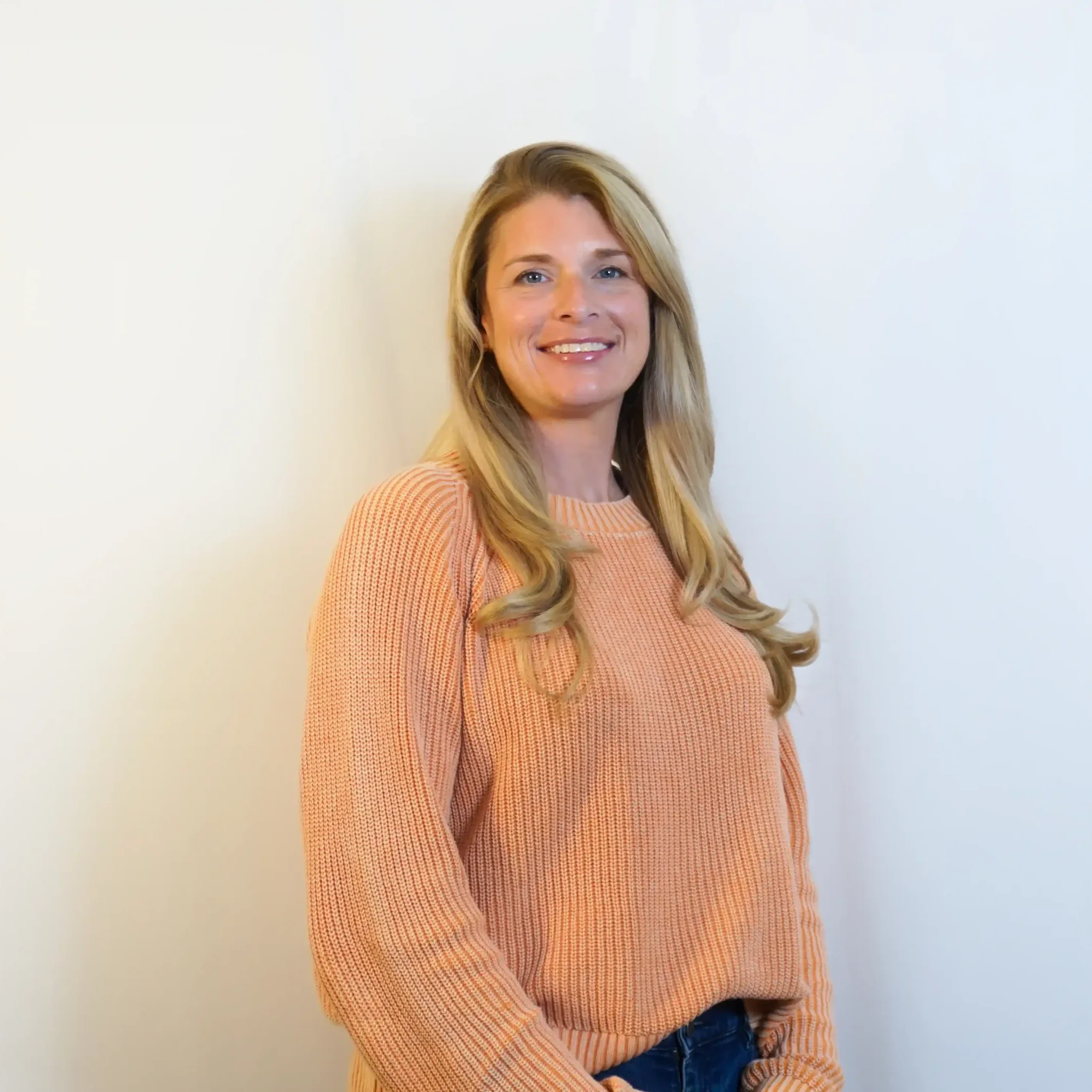 A blond haired woman in a peach colored sweater standing in front of a neutral back drop and smiling at the camera as she poises for a headshot as a public adjuster.