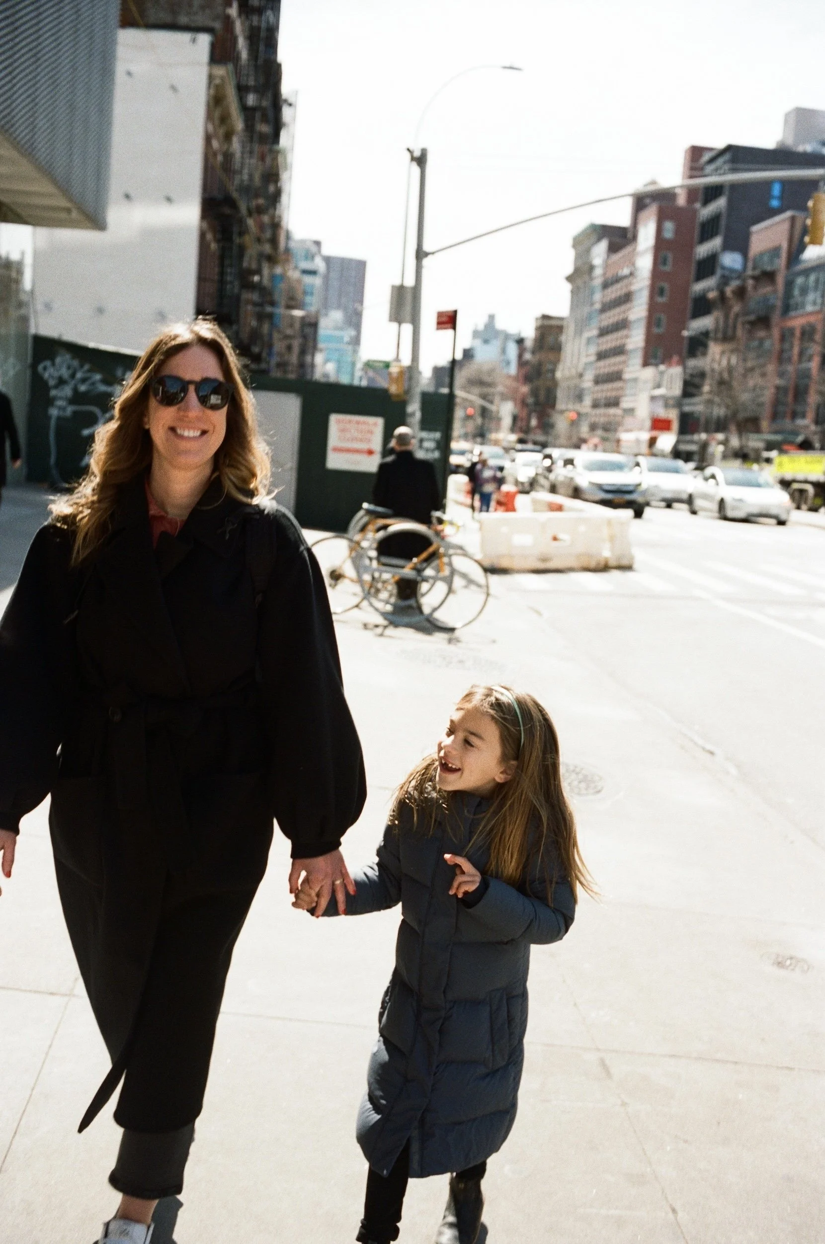 A woman and a young girl walking hand-in-hand on a city sidewalk with buildings, cars, and street activity in the background.