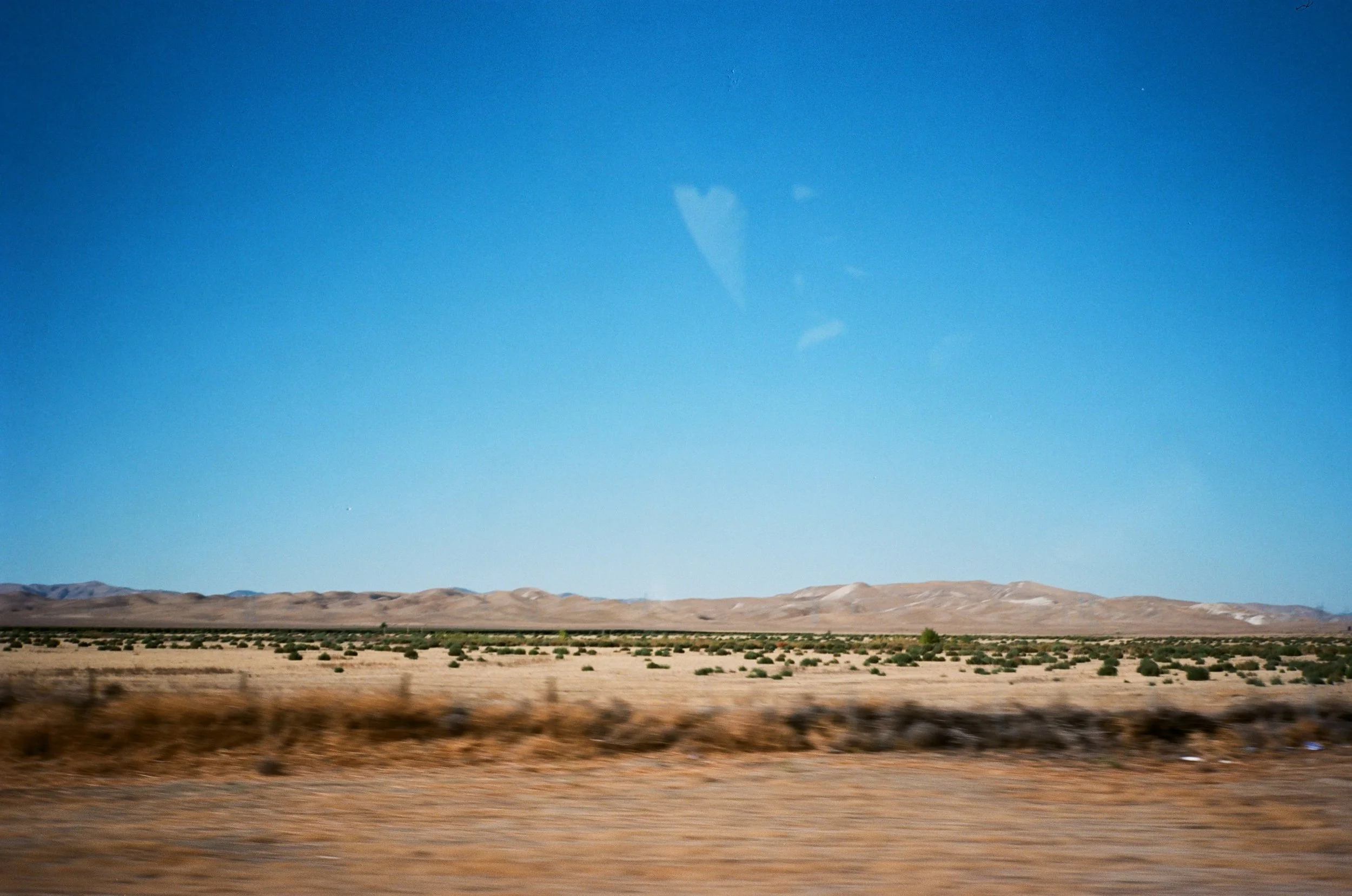 View of a barren desert landscape with distant hills under a clear blue sky, seen from a moving vehicle.