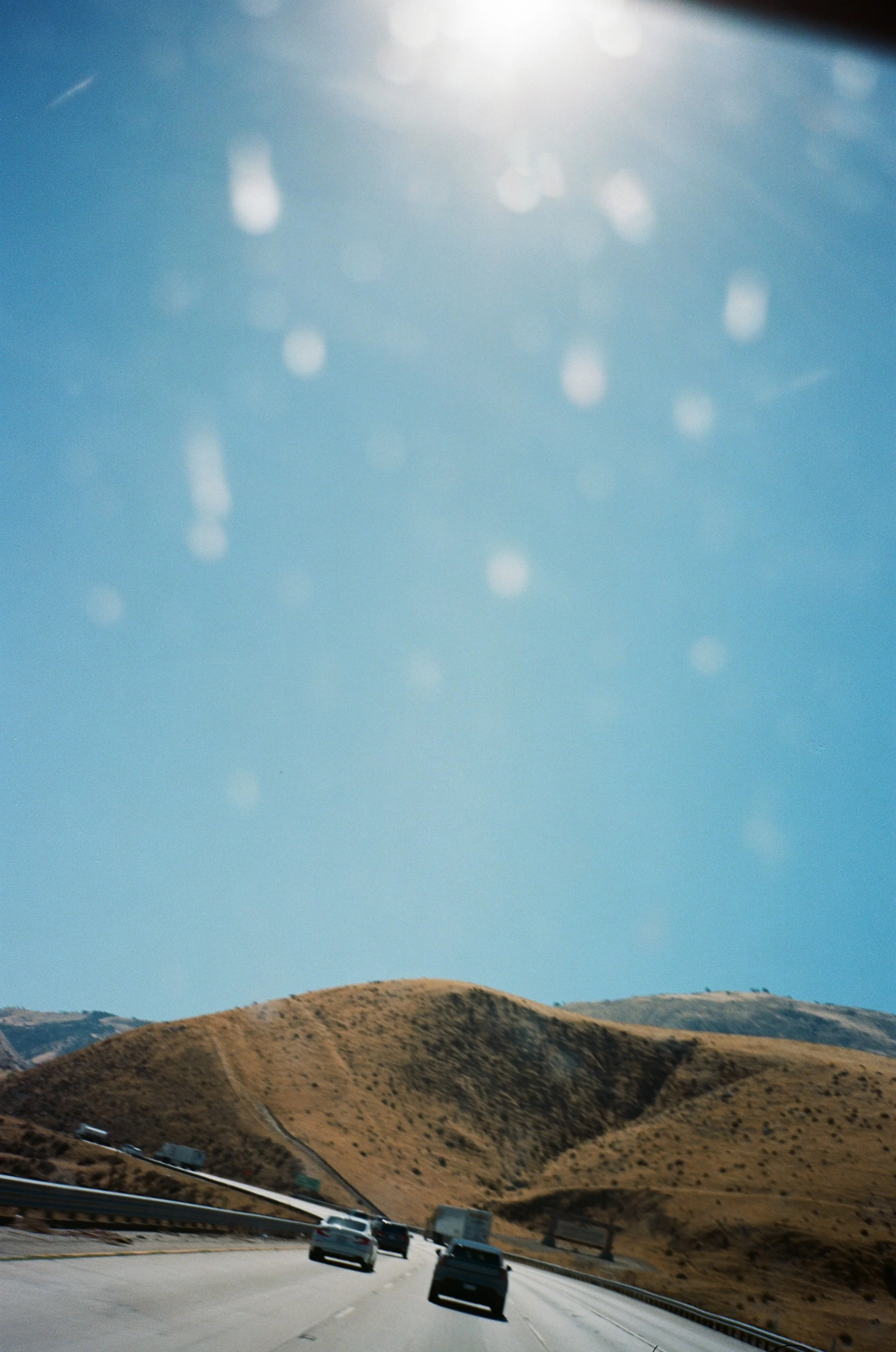 View of a highway with cars driving past dry, rolling hills under a clear blue sky.