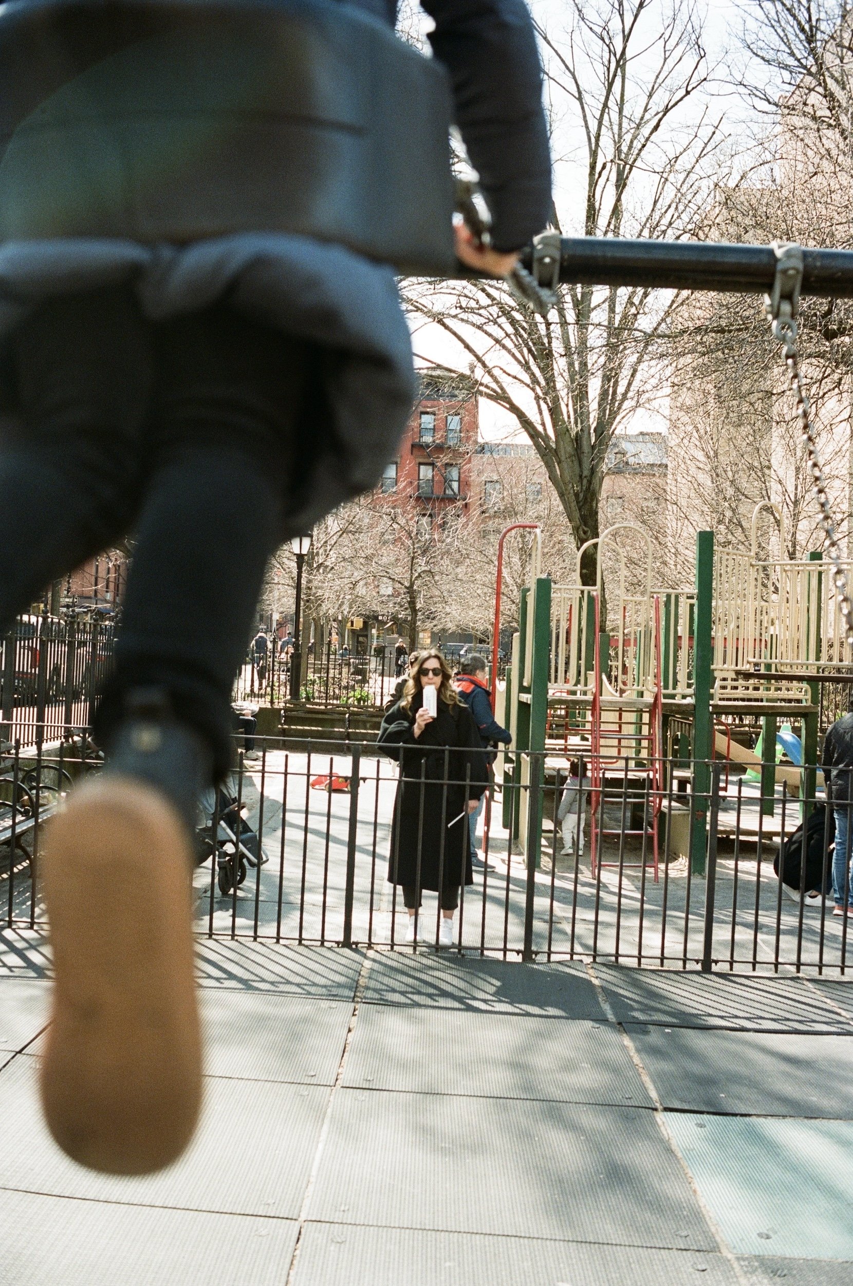 A woman in sunglasses taking a selfie in a playground with a gate, swing set, and trees in the background.