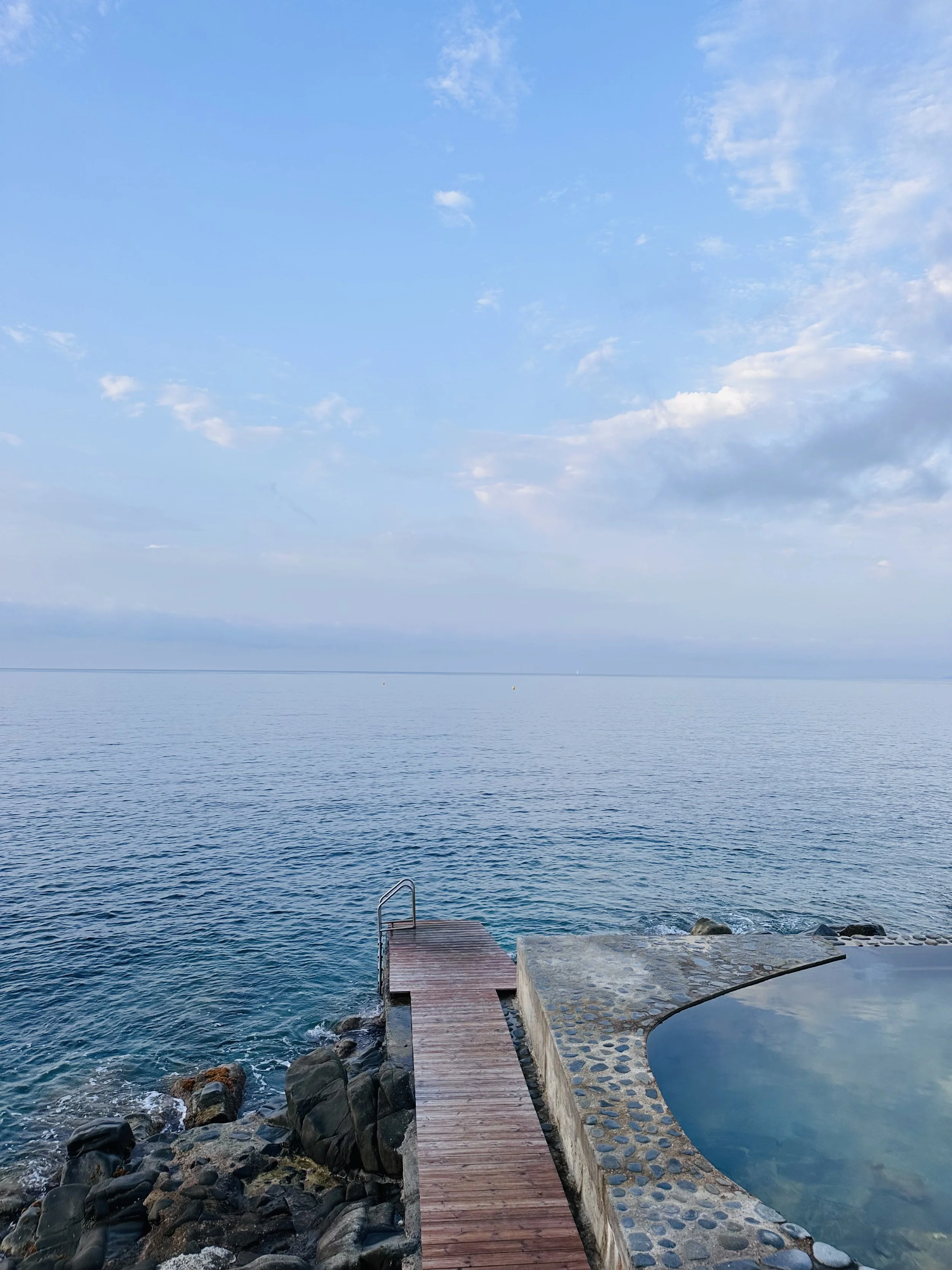 A serene view of the ocean with a clear blue sky and few scattered clouds, a small dock extending into the water, rocks along the shoreline, and a hot tub on the right side of the image.