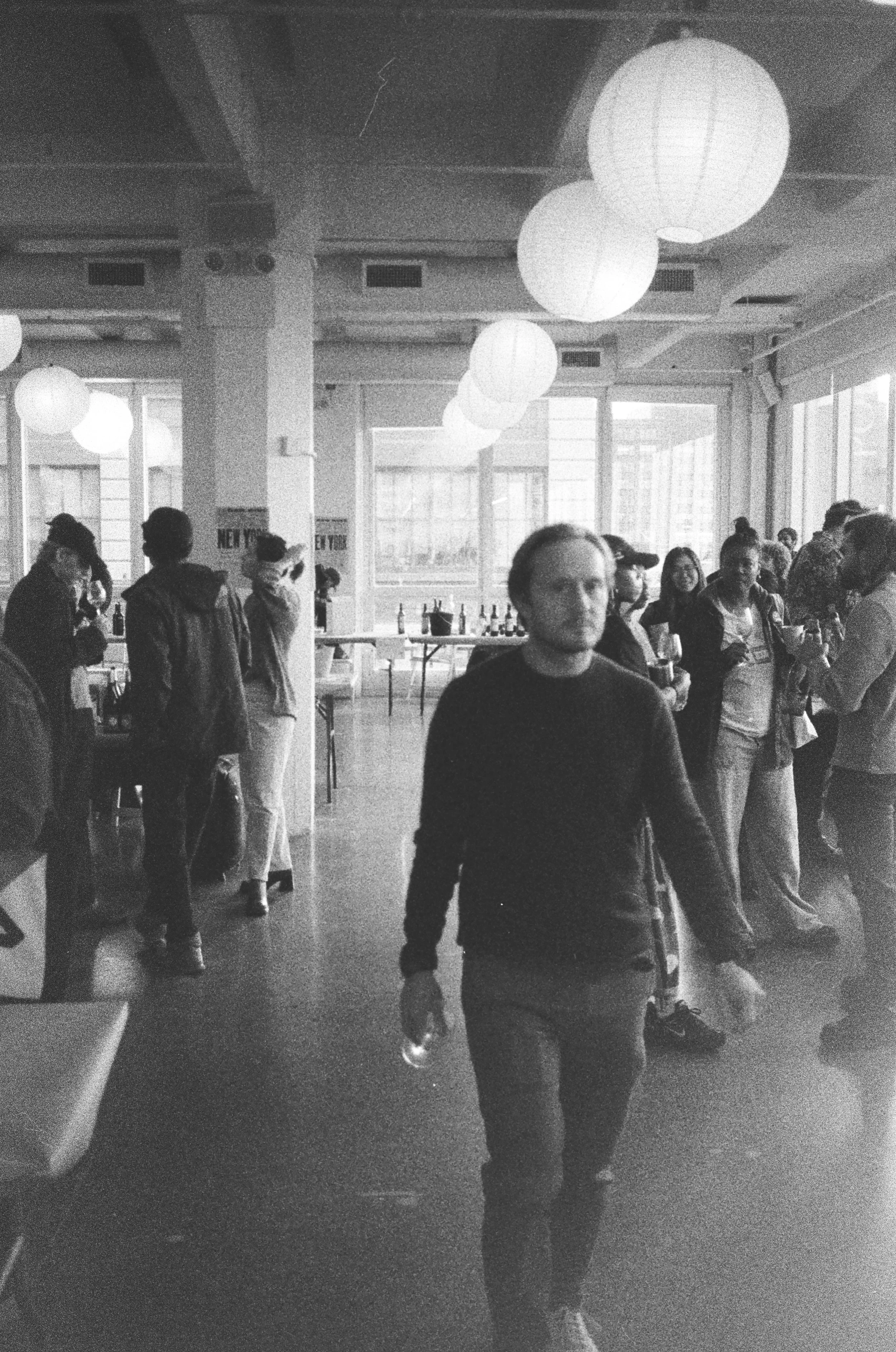 People socializing at an indoor gathering with large paper lantern lights hanging from the ceiling, some holding drinks, in a bright room with large windows.