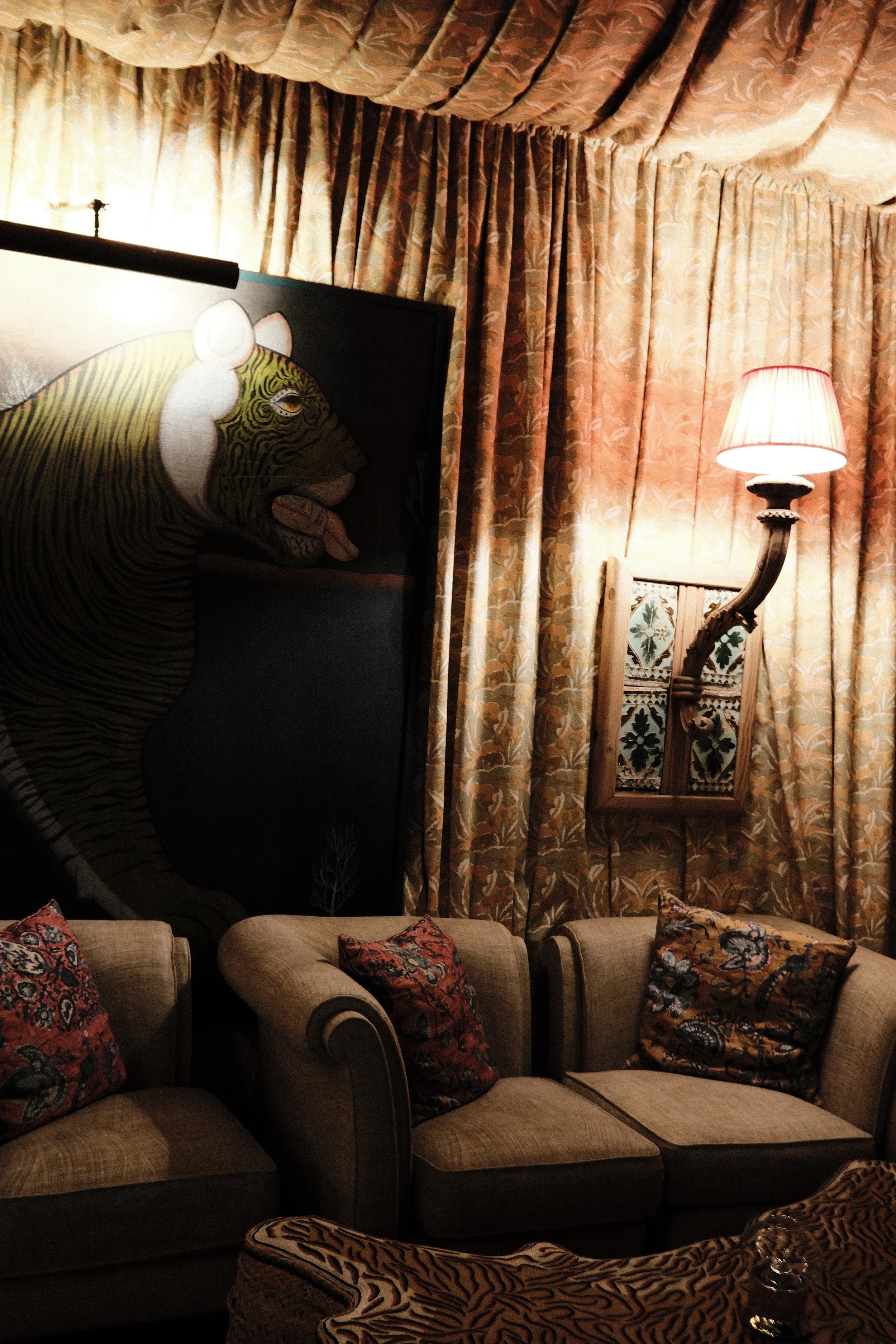 Cozy room with patterned curtains, a wall-mounted lamp, a mirror, a large tiger painting, and comfortable chairs with decorative pillows.