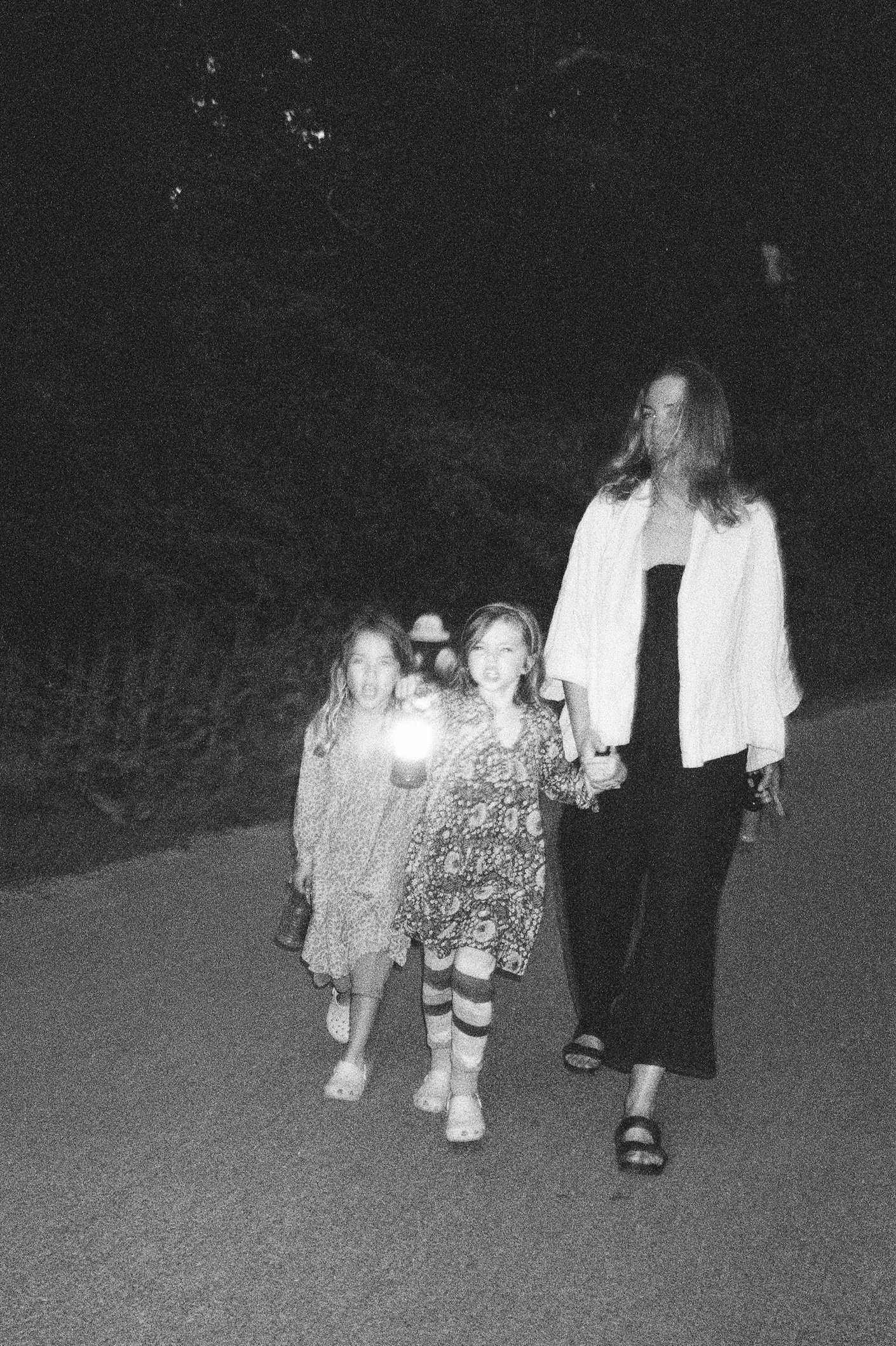 A woman walking hand-in-hand with two young girls at night on a paved path, with trees in the background. The woman is wearing a light-colored jacket, black pants, and sandals. The girls are dressed in patterned dresses and socks, and one is holding 