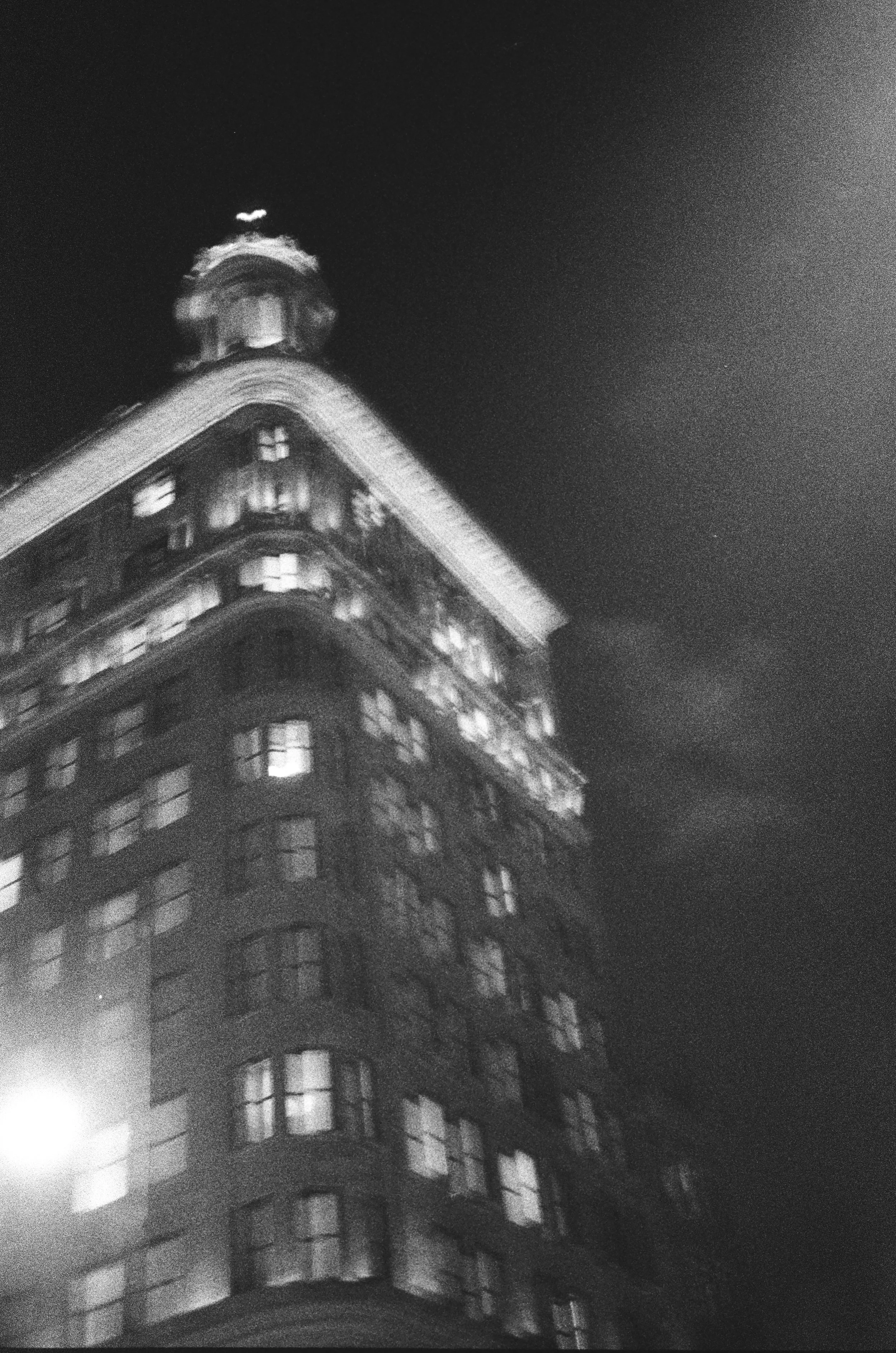 A black-and-white night photo of a tall, historic building with illuminated windows and a rounded corner with a tower on top, against a dark sky.