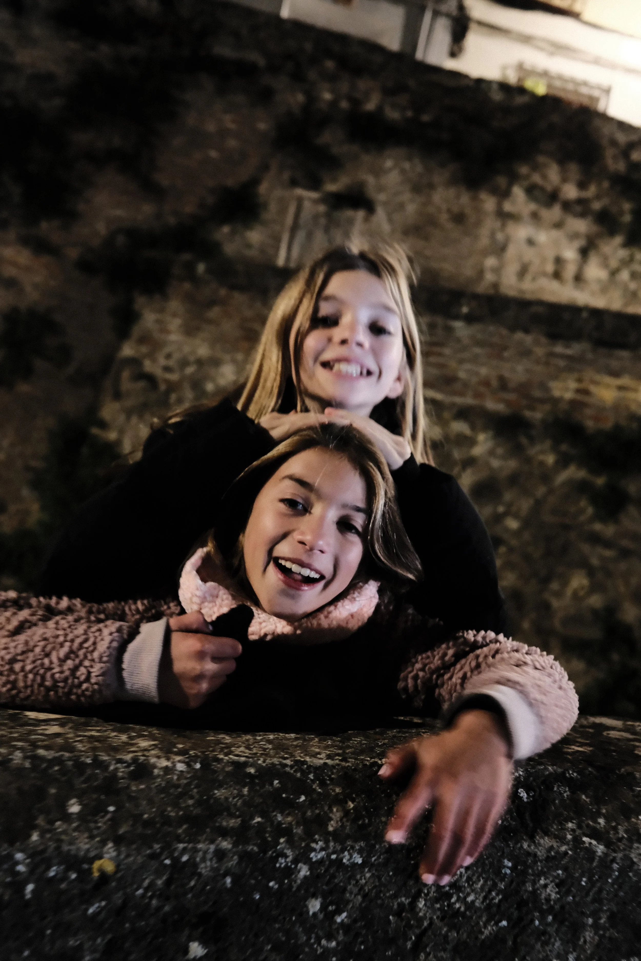 Two girls smiling and playing on a large rock at night, with one girl lying on her stomach in front and the other girl leaning on her back, both making playful expressions.