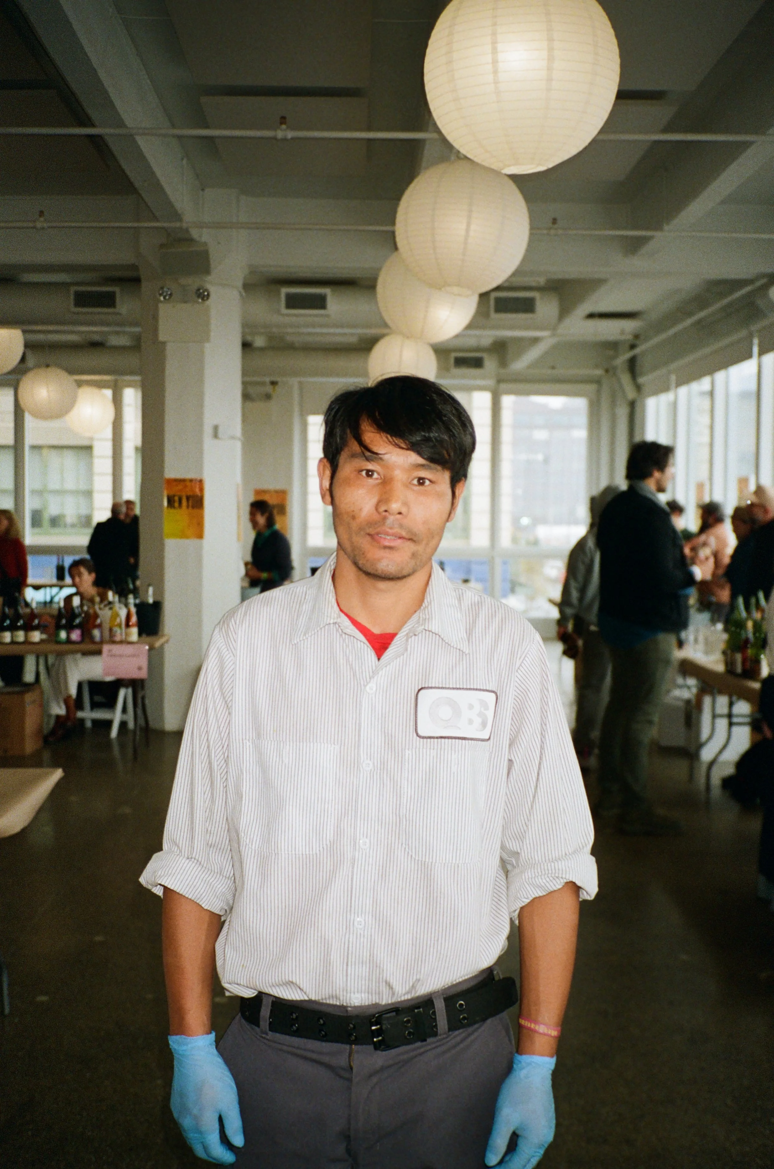 A man standing in a well-lit interior space with hanging round paper lanterns, many people in the background, some preparing food, some socializing, large windows letting in natural light.