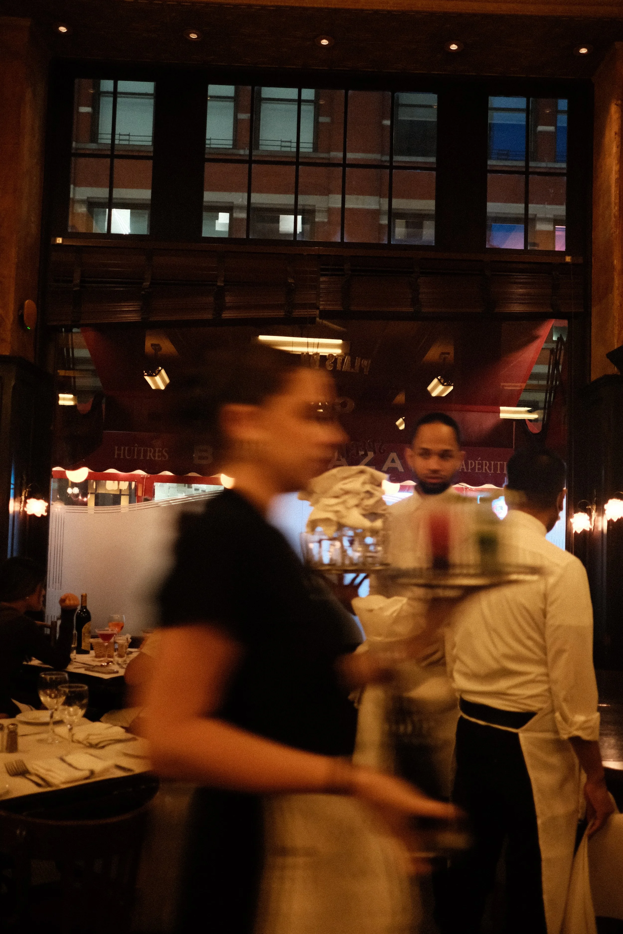 A busy restaurant with waitstaff and diners, during evening hours, with large windows and warm interior lighting.