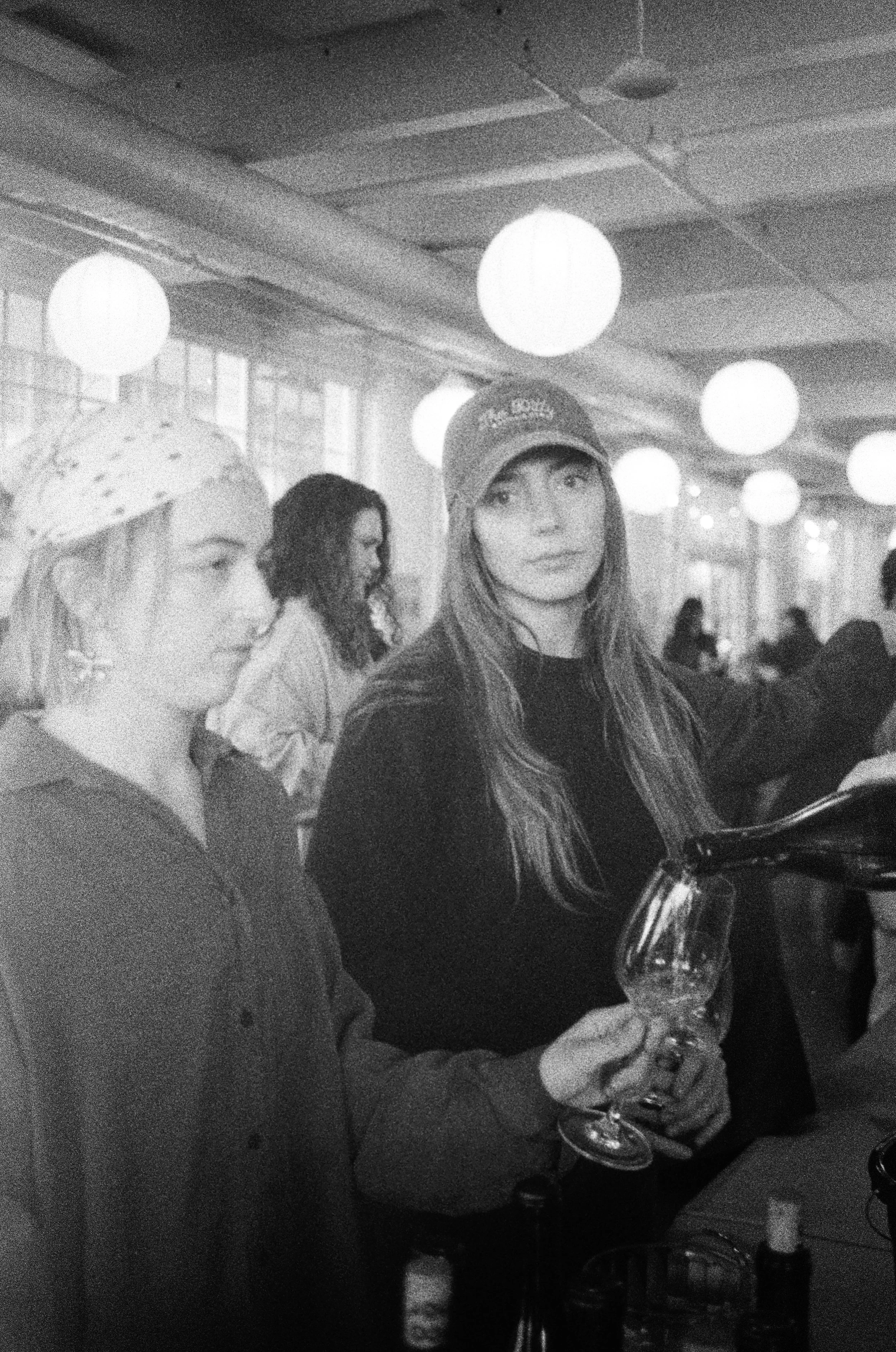 Two young women standing at a bar in a dimly lit restaurant or cafe, one wearing a bandana and the other wearing a cap, with drinks in their hands, surrounded by hanging round light fixtures and other people in the background.