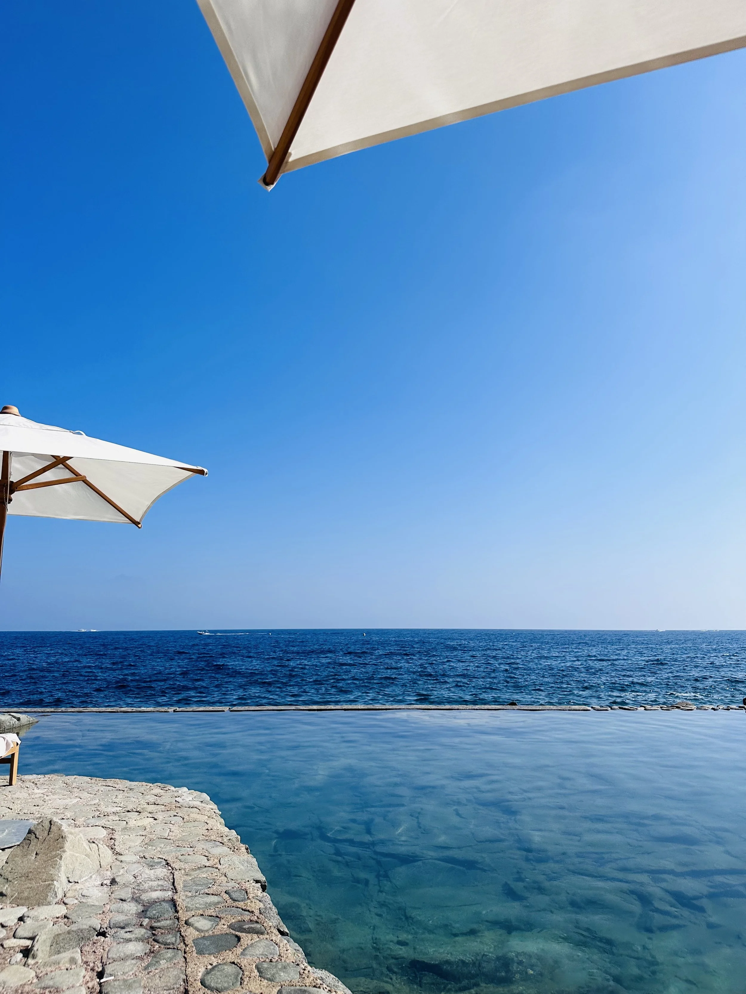 View of the ocean with a stone pier and two large white umbrellas, bright blue sky, and distant boats.