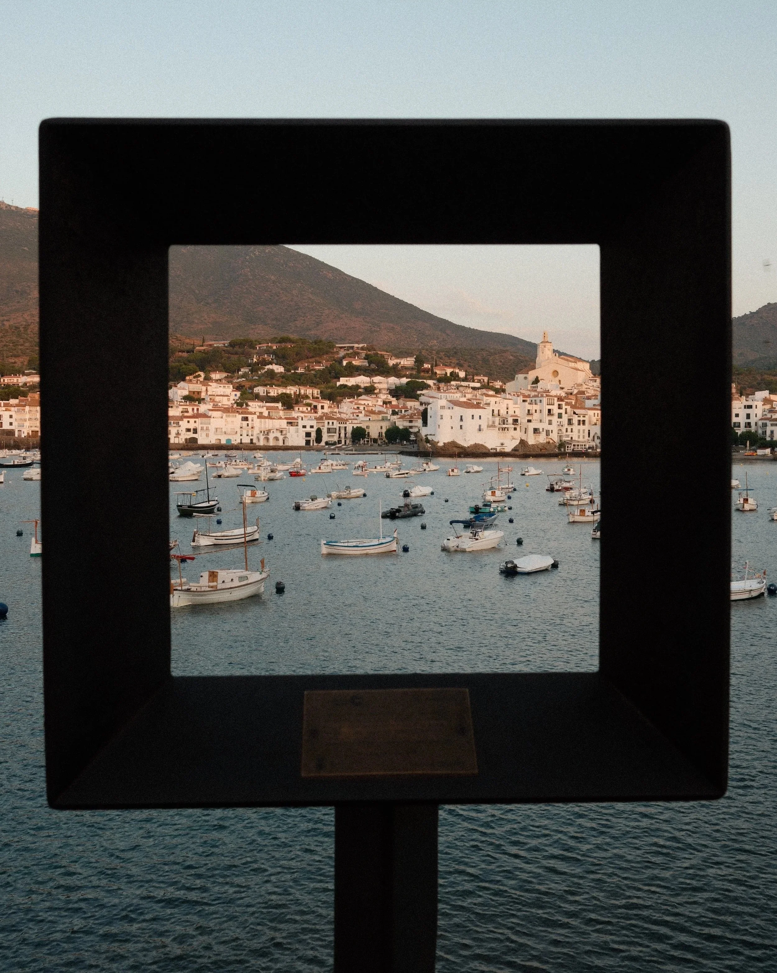 View of a harbor with small boats, seen through a square black frame, with white buildings and hills in the background.