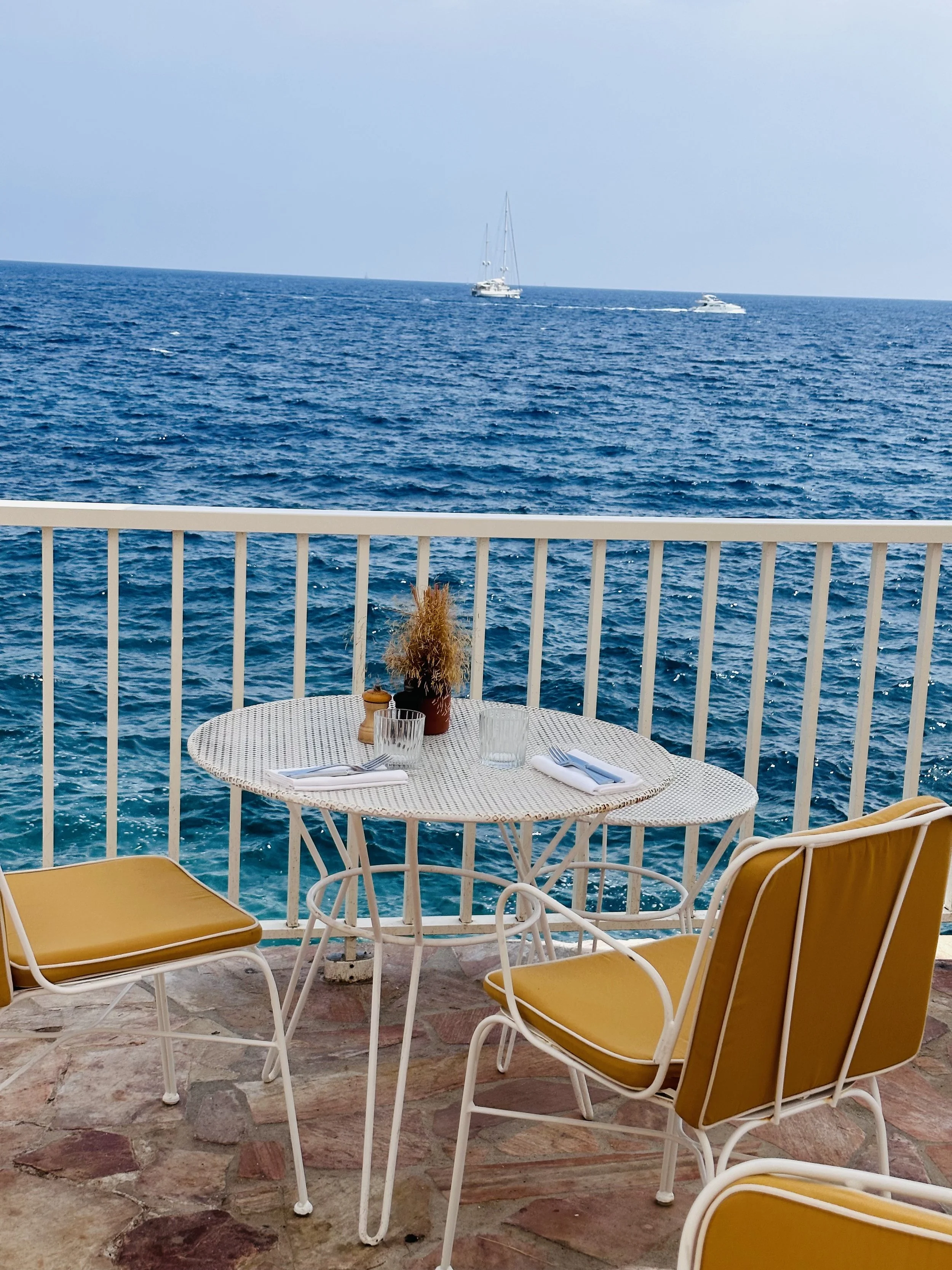 A seaside outdoor dining area with a round table set with two glasses, cutlery, and a plant in a pot, overlooking the ocean with boats sailing in the distance.