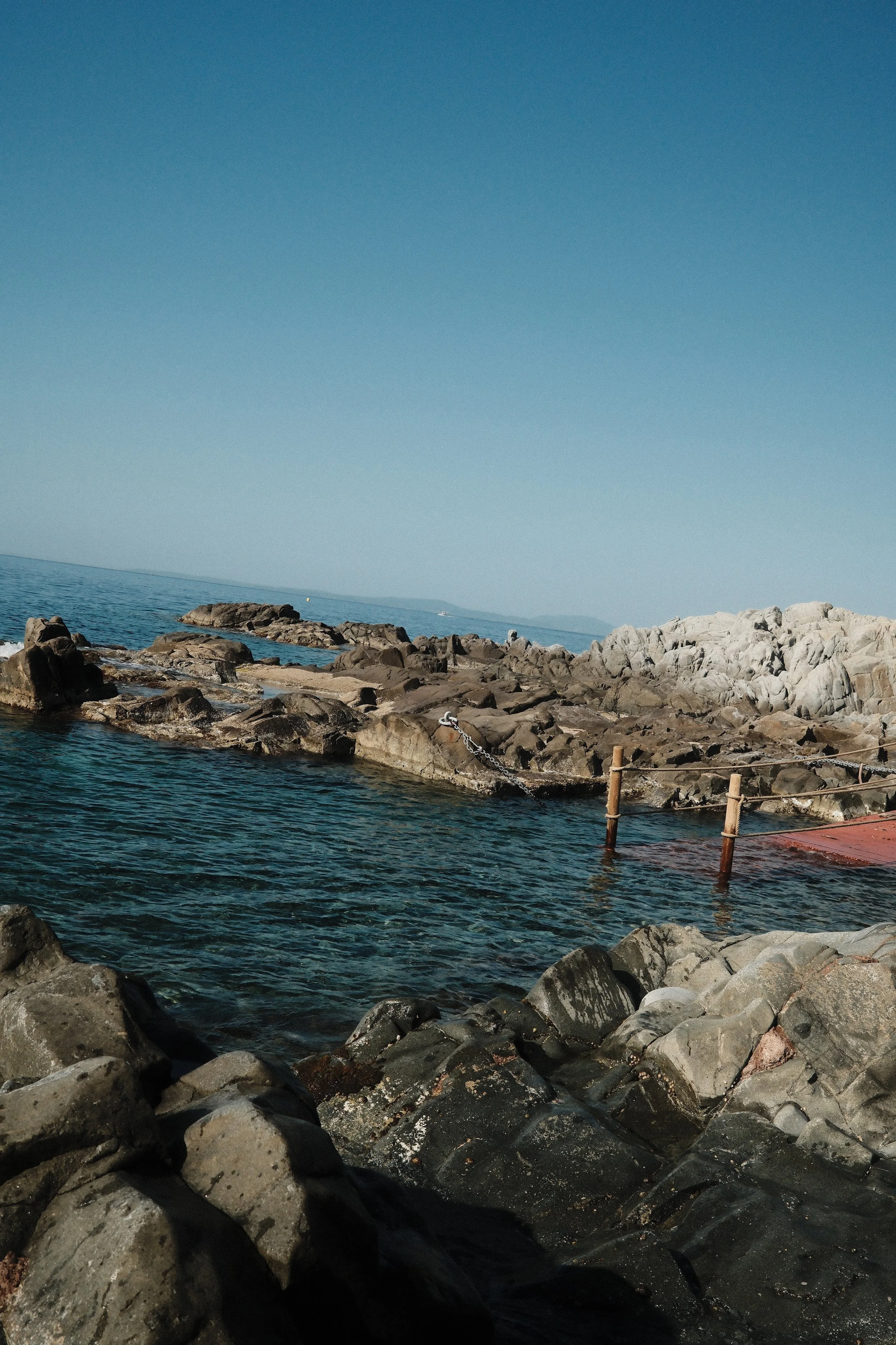 A rocky shoreline with rocks extending into the ocean, a clear blue sky, and distant landforms on the horizon.