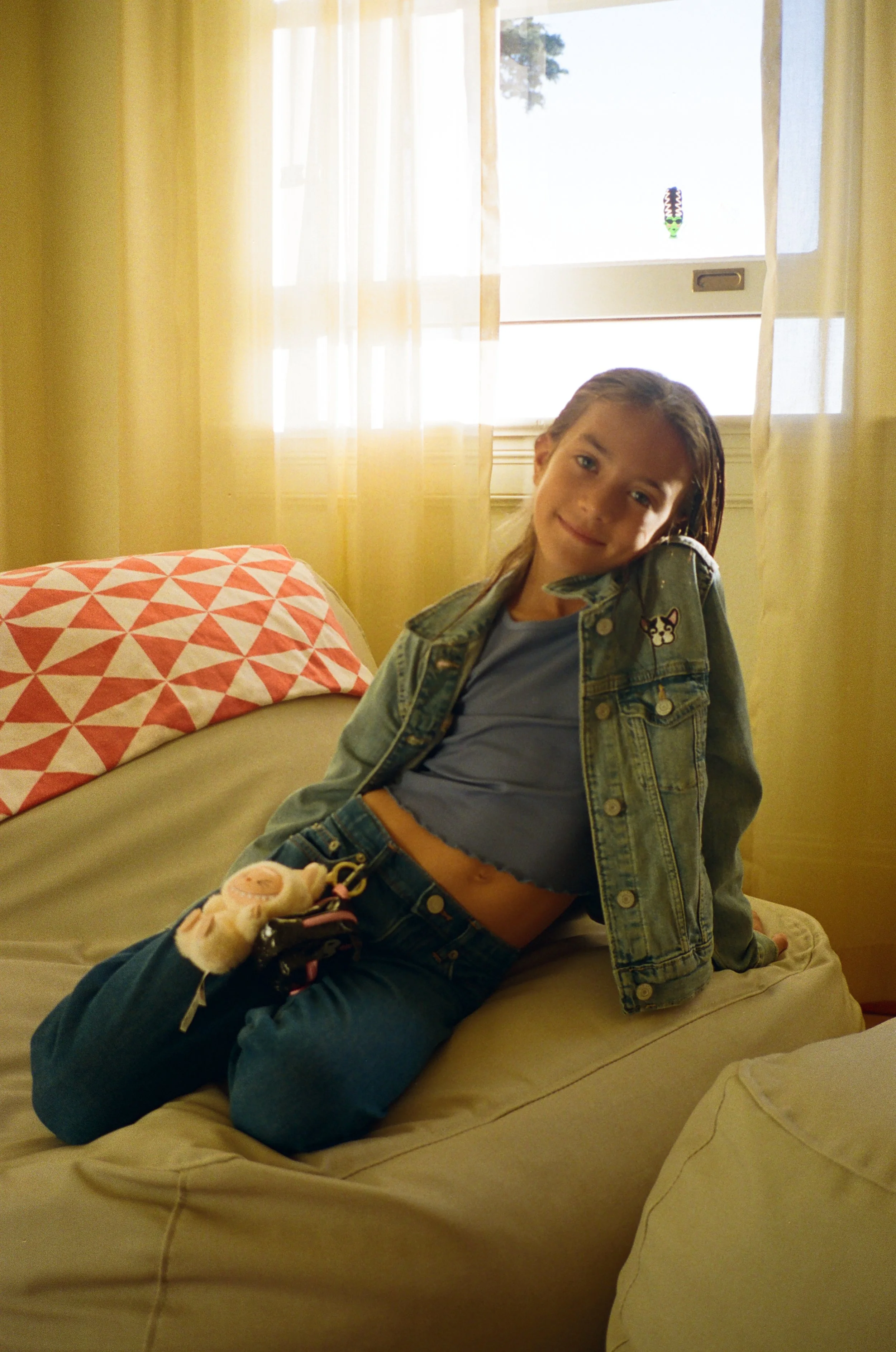 A girl with long brown hair, wearing a denim jacket and blue shirt, sitting on a beige sofa with a patterned pillow, in a room with yellow walls and a window with light-colored curtains.