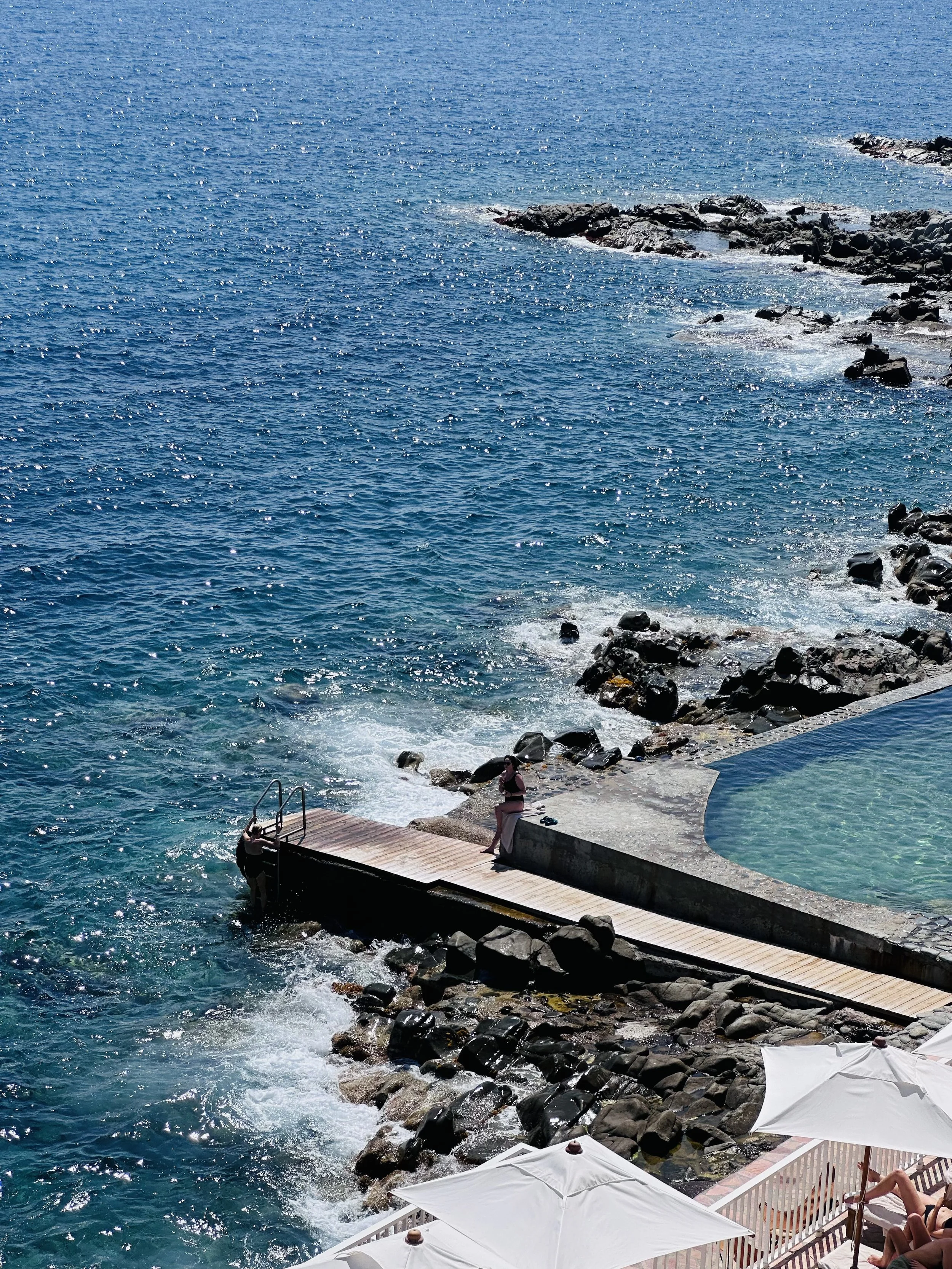 A seaside scene with clear blue water, a rocky shore, a wooden dock, and a swimming pool with people relaxing under white umbrellas.