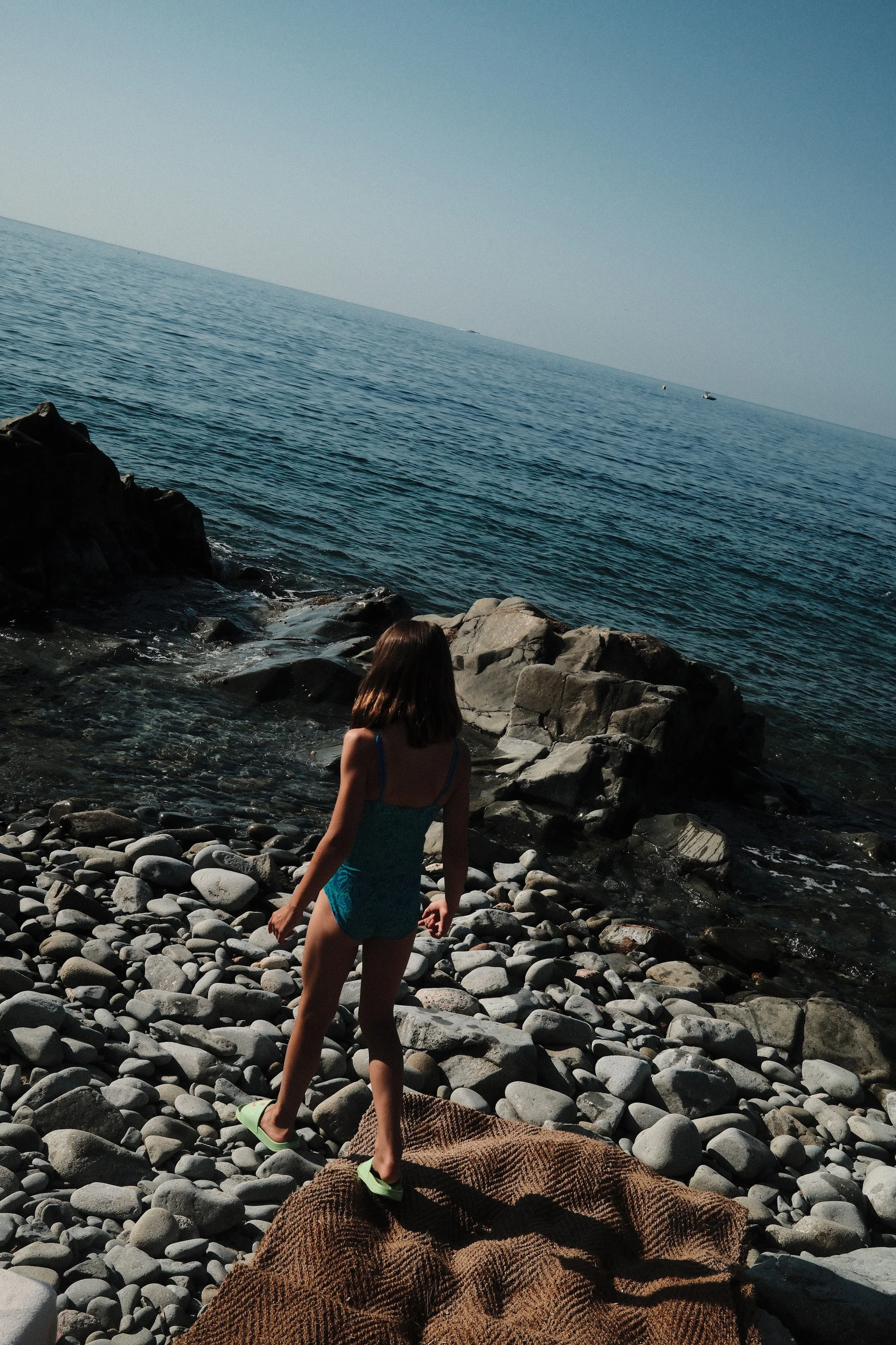 A girl in a blue swimsuit and green flip flops walking on a rocky beach near the ocean with rocks and waves, and a person lying on a brown towel in the foreground.