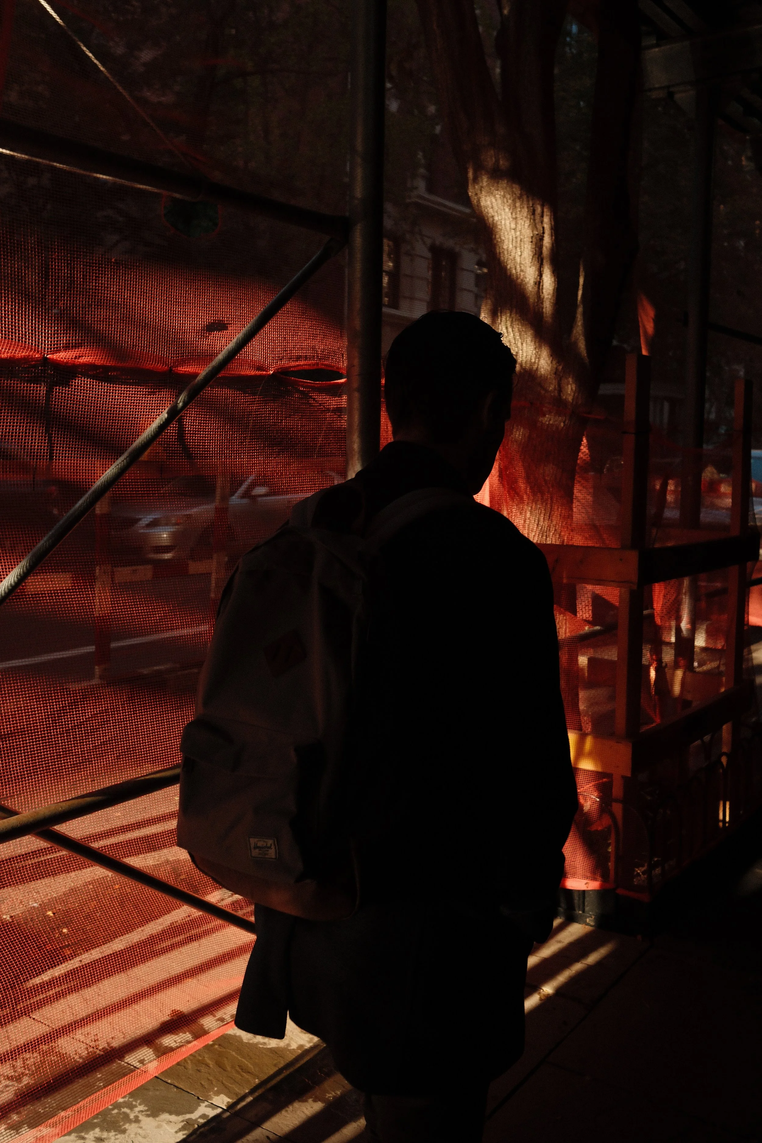 Silhouette of a person standing outdoors near a construction barrier with red mesh, in front of a tree and building shadows, during sunset or dusk.