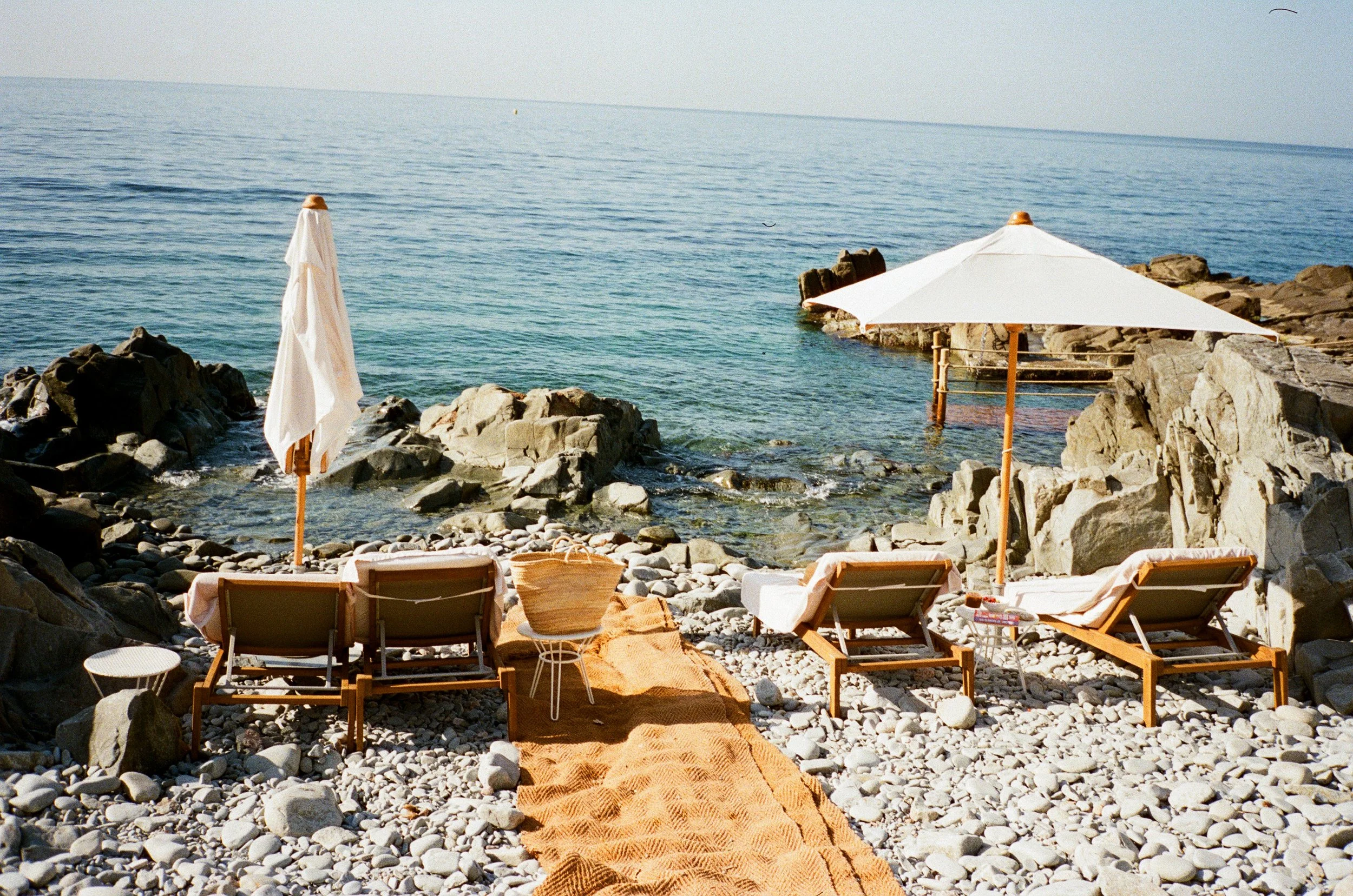 Beach scene with lounge chairs, white umbrellas, rocky shoreline, and calm ocean water.