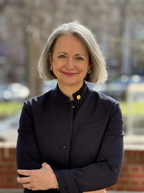 Dr. Adriana Kipper-Williams. A smiling middle-aged woman with shoulder-length gray hair, wearing a black jacket, standing outdoors with a blurred background of trees and cars.