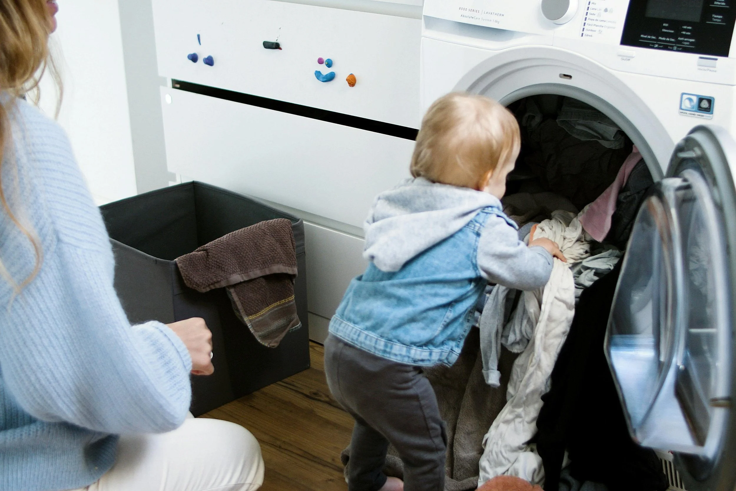 Parent and young child loading laundry into a washing machine, representing the physical and mental labor mothers carry at home.